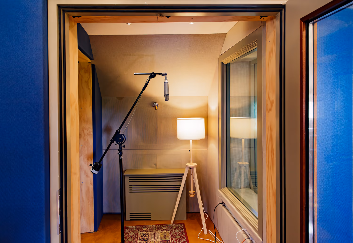 Studio 1 isolation booth seen through an open doorway, condenser mic on a boom stand beside a tripod floor lamp