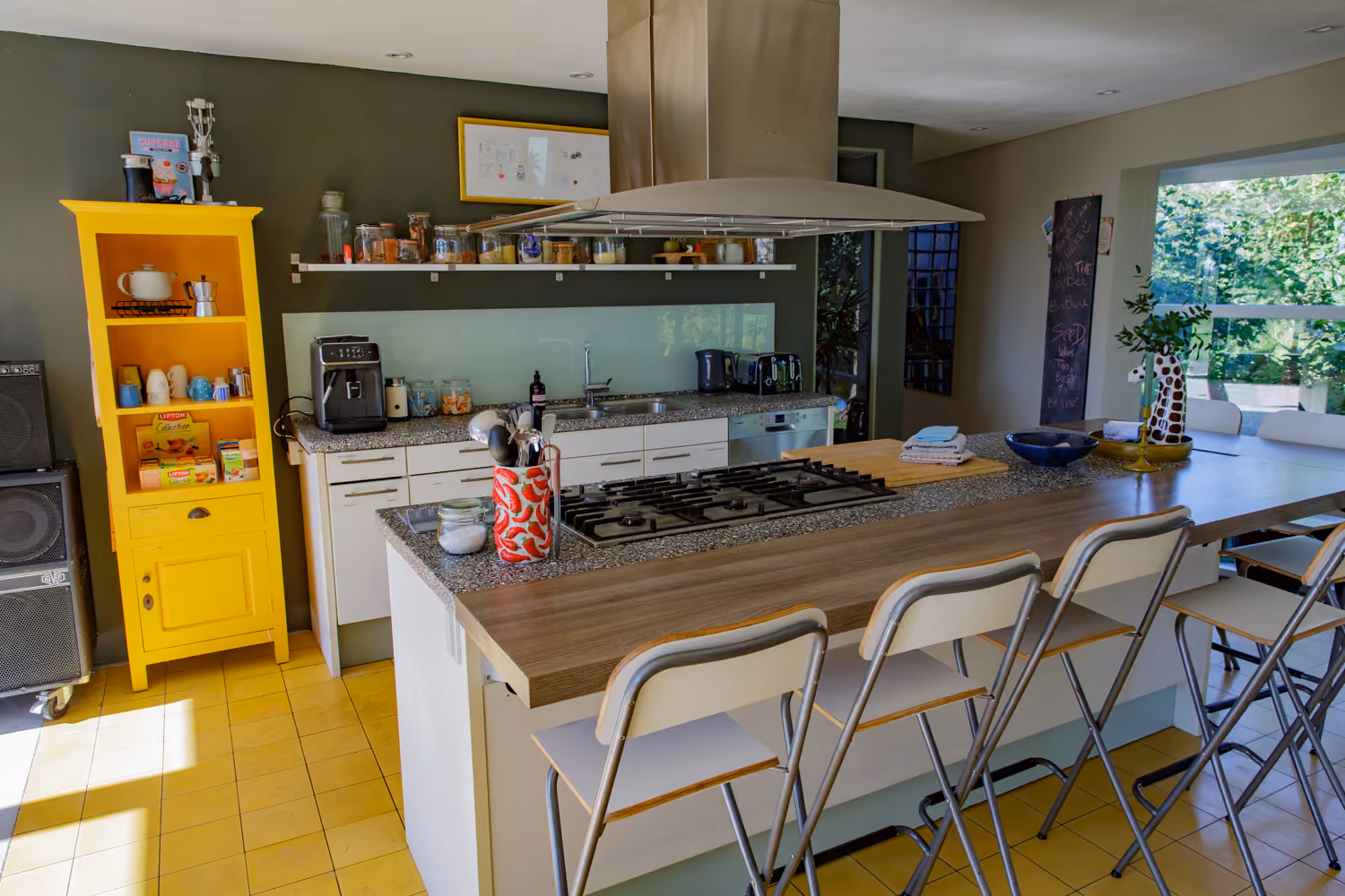 Guest area kitchen with gas hob island, folding bar stools, yellow cupboard and an open shelf of glass storage jars