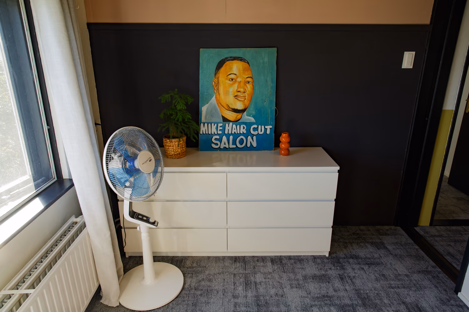 White dresser with a hand-painted Mike Hair Cut Salon sign, potted plant and standing fan against a dark wall