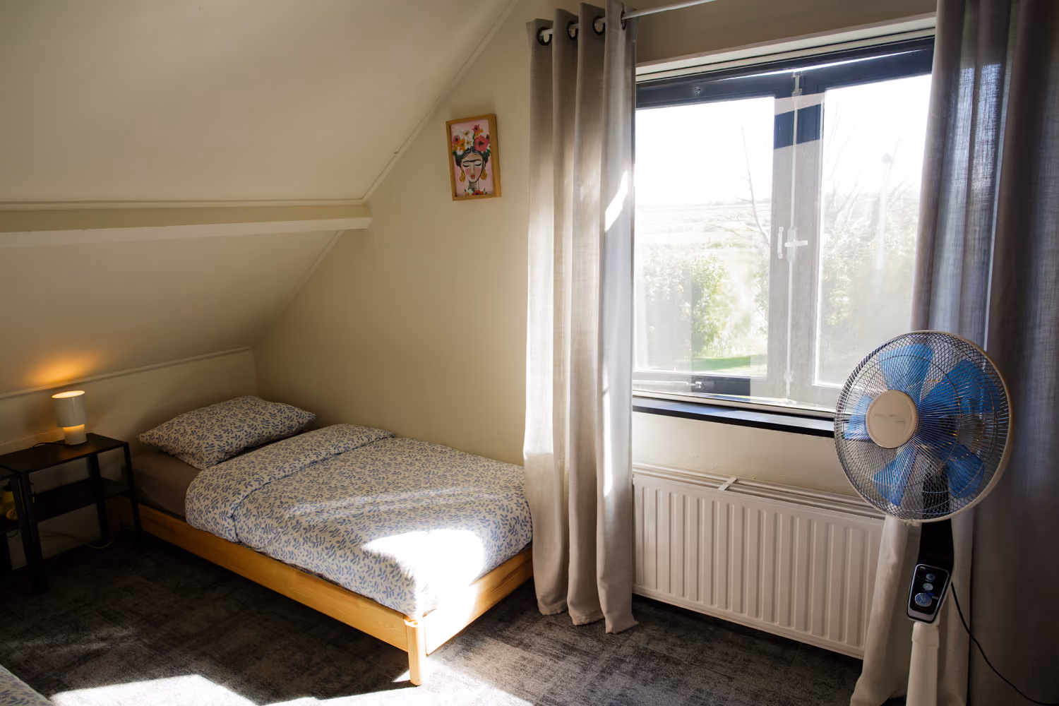 Bedroom upstairs with a single pine bed, framed floral portrait, standing fan and a window overlooking fields