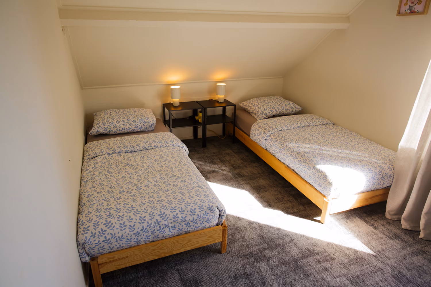 Bedroom upstairs with two single pine beds in blue leaf-print bedding, white lamps on black nightstands and a sloped ceiling