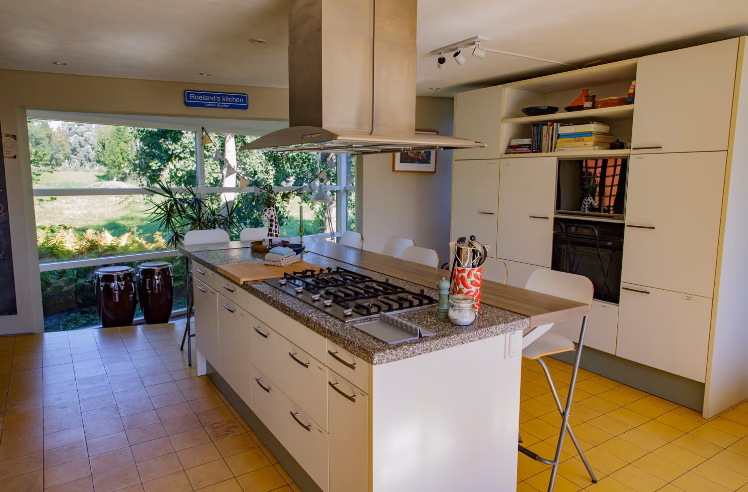 Guest area kitchen island with gas hob and steel extractor hood, conga drums by a big window onto the garden