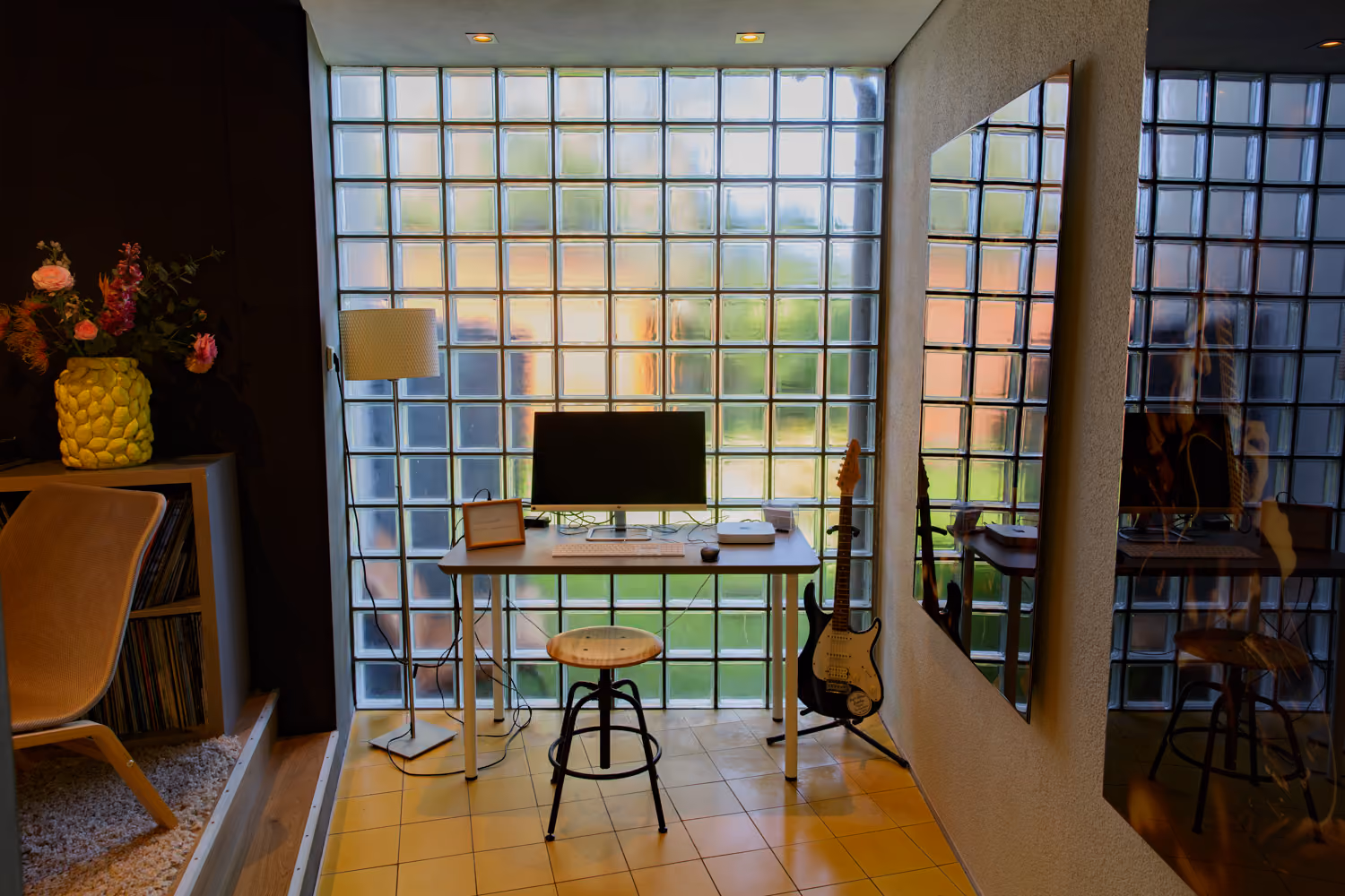 Small desk with a monitor and Mac mini, a wooden stool and an electric guitar in front of a glass-block wall
