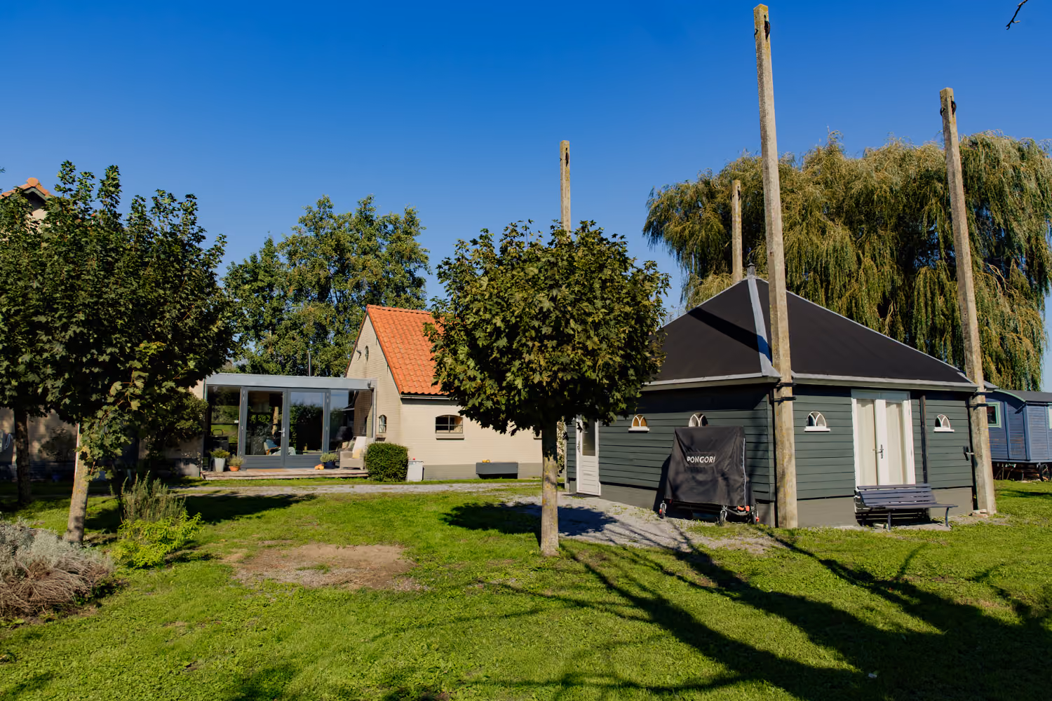 Sunny farm lawn with a brick farmhouse and glass extension, a dark green timber building and a weeping willow