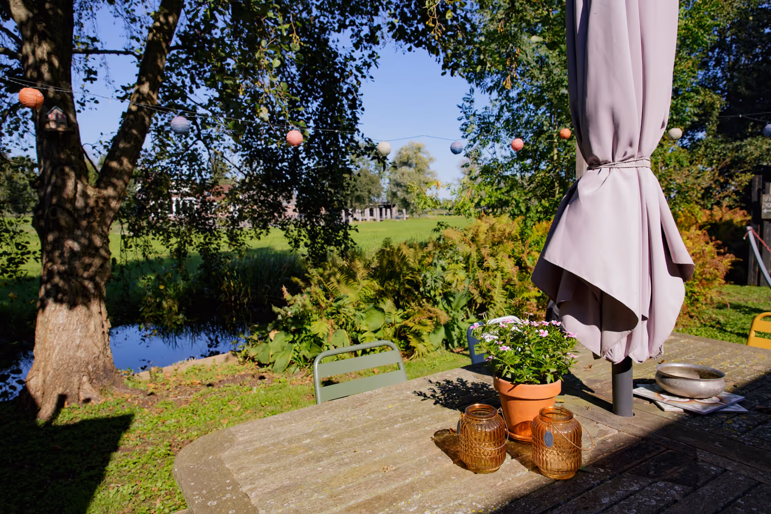 Back garden terrace table with pink flowers and amber lanterns, paper lanterns over a pond and meadow beyond