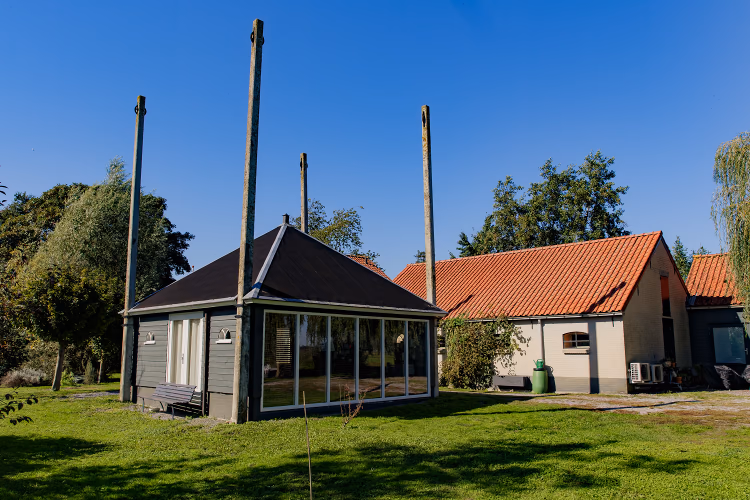 Dark timber garden pavilion with a glass wall and tall corner posts beside the cream brick barn with orange roof tiles