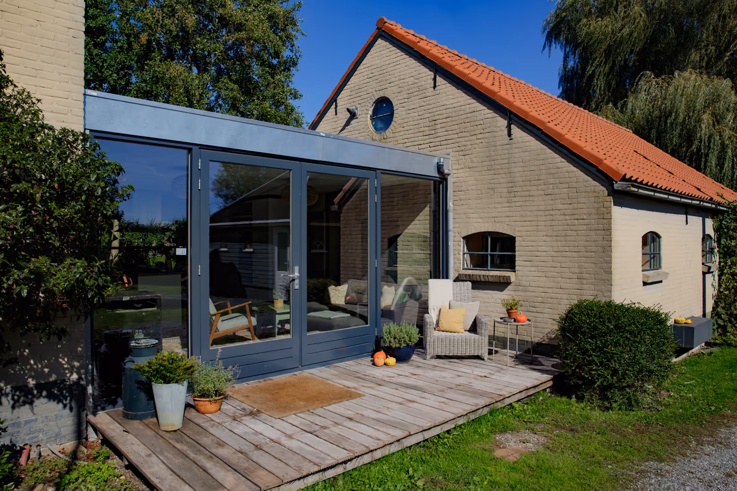 Wooden deck and glass-walled lounge with a sofa and armchair inside, beside a red-tiled brick farm building