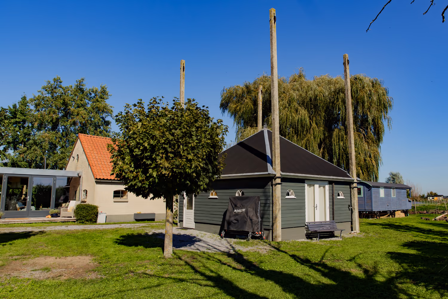 Dark green timber building with small arched windows and concrete poles, a blue caravan and a weeping willow