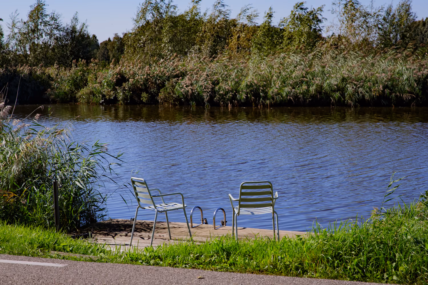 Two pale green chairs and a swim ladder on a small wooden jetty on the Amstel river, reeds on the far bank