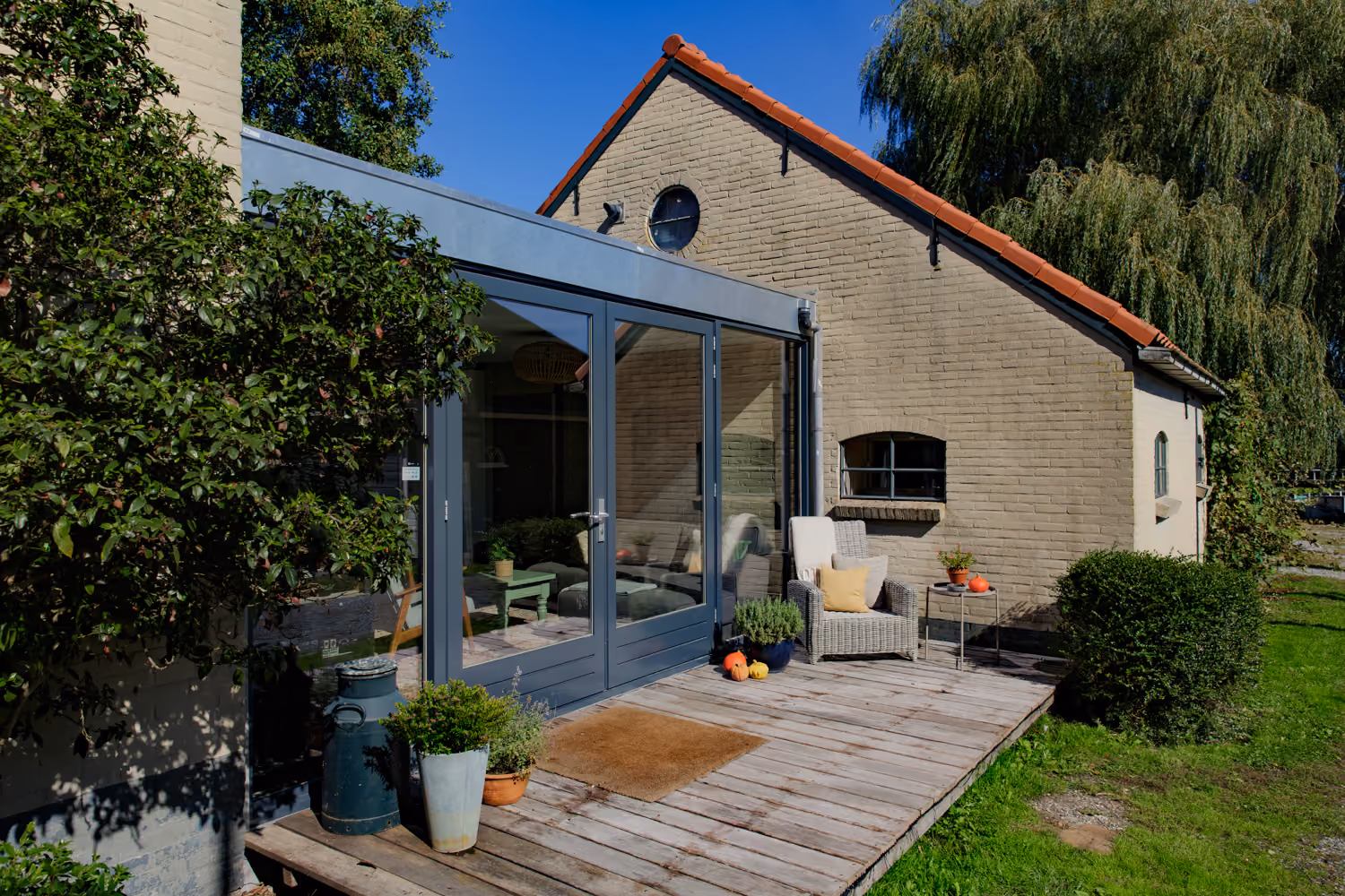 Glass-fronted lounge extension and wooden deck with a milk churn and wicker armchair beside a red-tiled brick gable