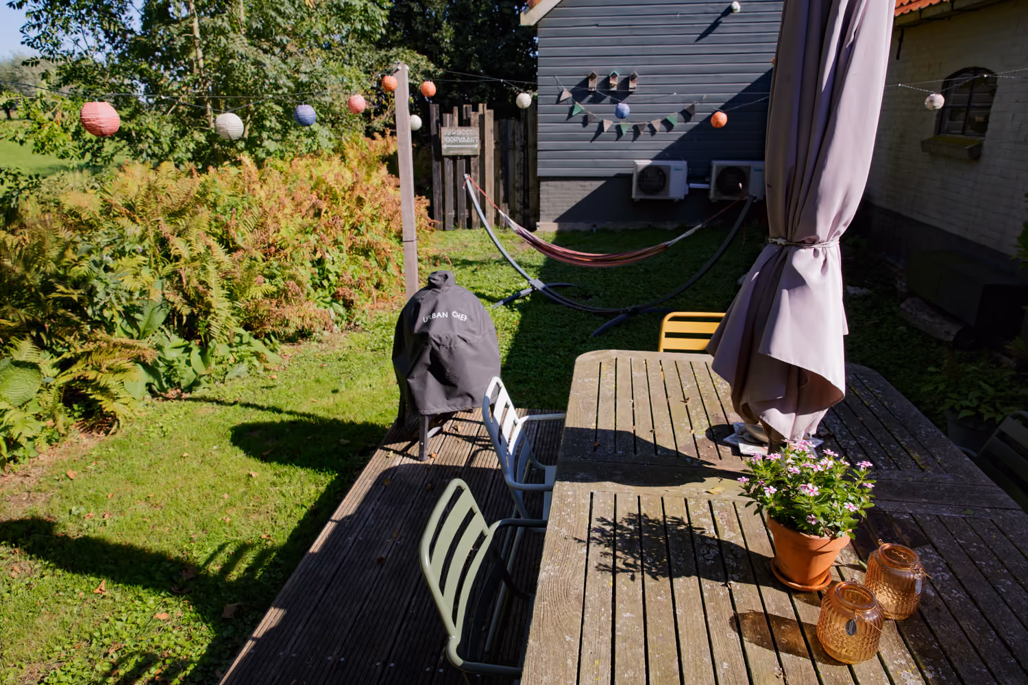 Back garden terrace with a wooden table, covered barbecue and a hammock on the lawn under strings of paper lanterns