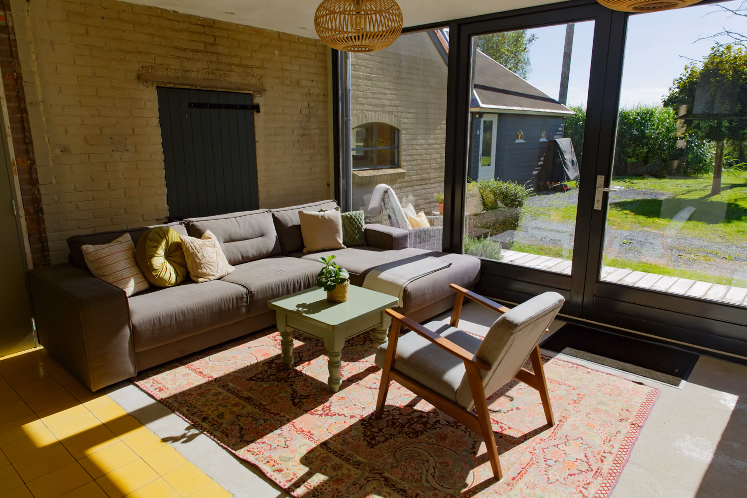 Guest area chill corner with a corner sofa, green coffee table and wooden armchair on a Persian rug by tall windows