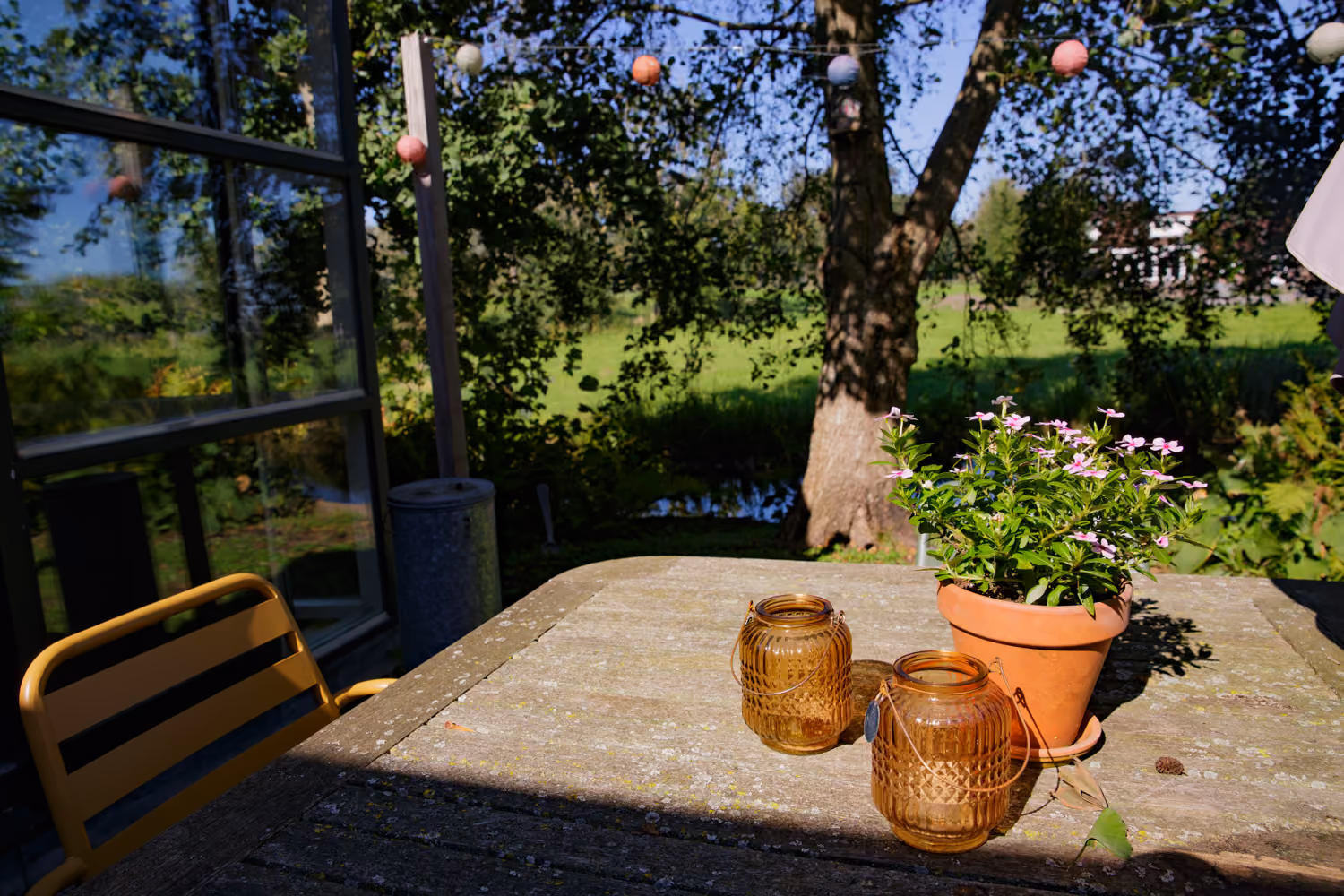 Back garden terrace table with amber glass lanterns and pink potted flowers, paper lanterns strung in the tree
