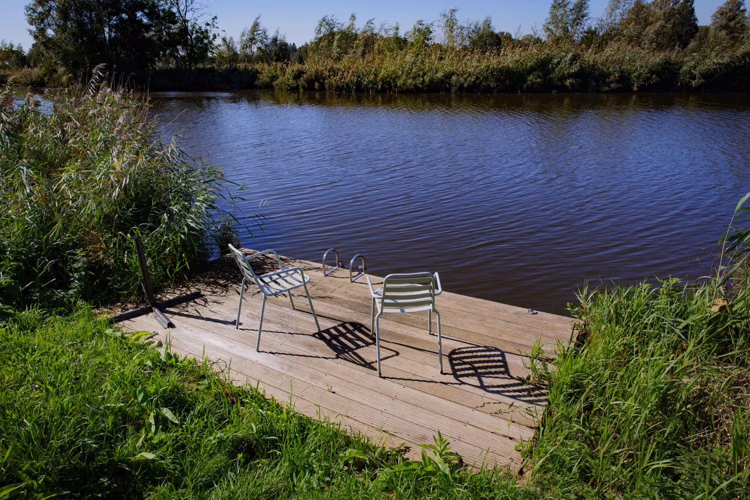 Wooden jetty on the Amstel with two pale green metal chairs and a swim ladder, reeds along both banks