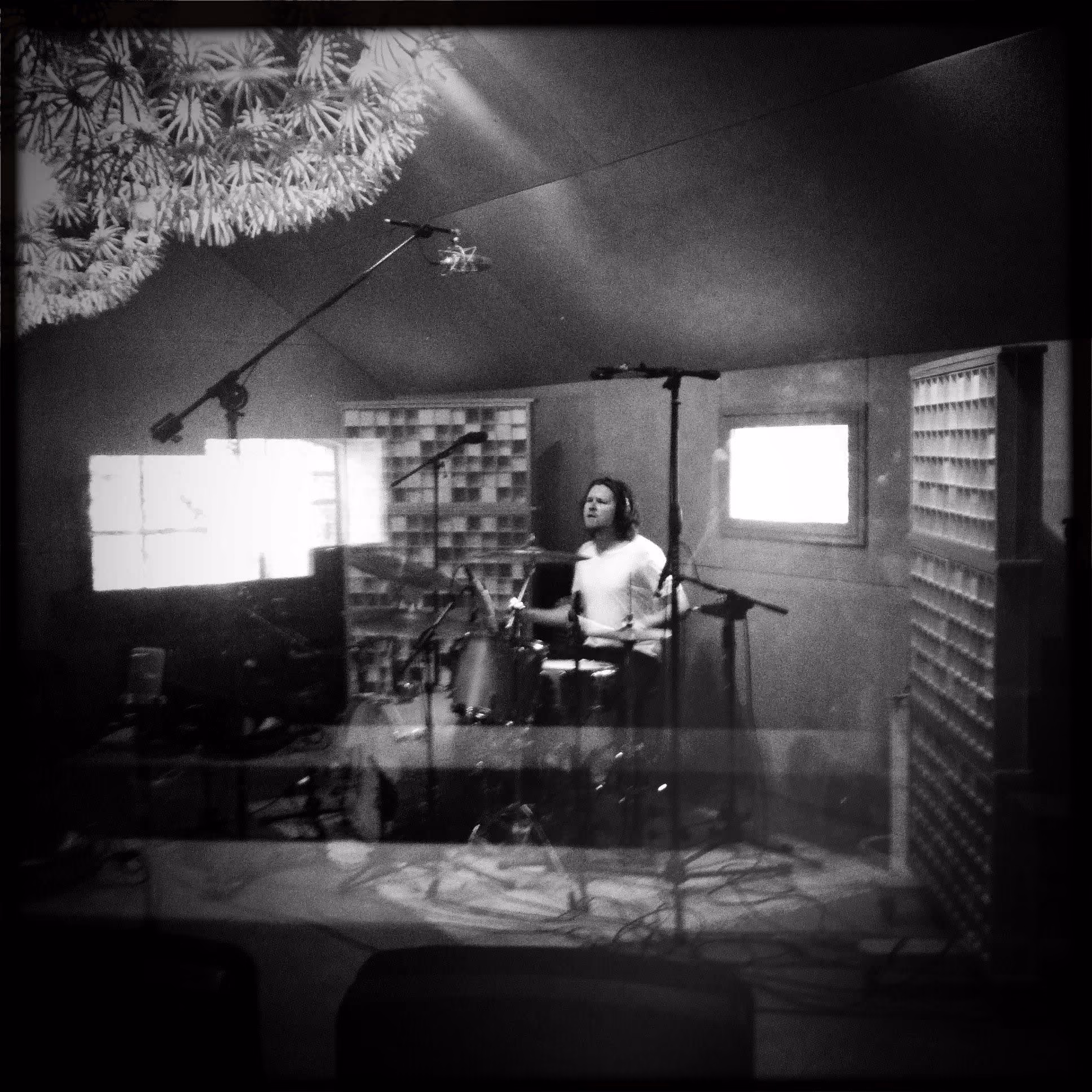 Black and white view through the glass of a drummer recording in the Studio 1 live room with overhead mics