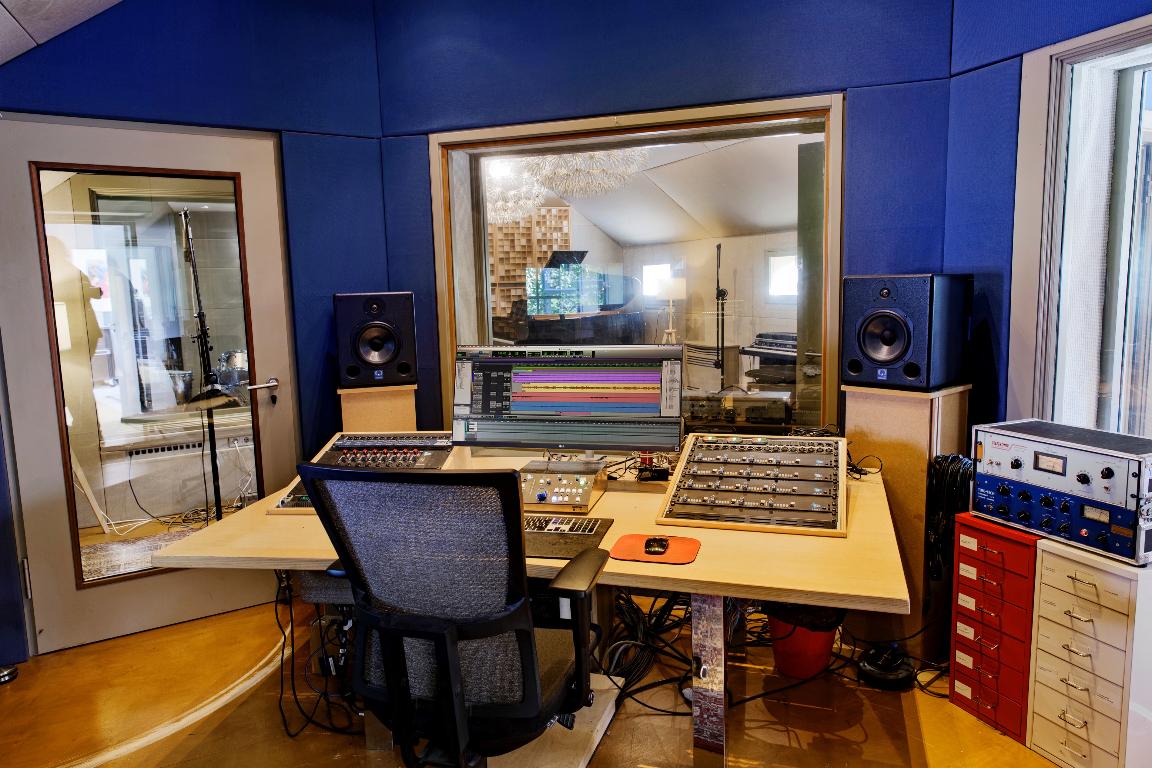 Studio 1 control room desk with a monitor, two studio speakers, an API preamp rack and outboard gear, and a window into the live room with the grand piano