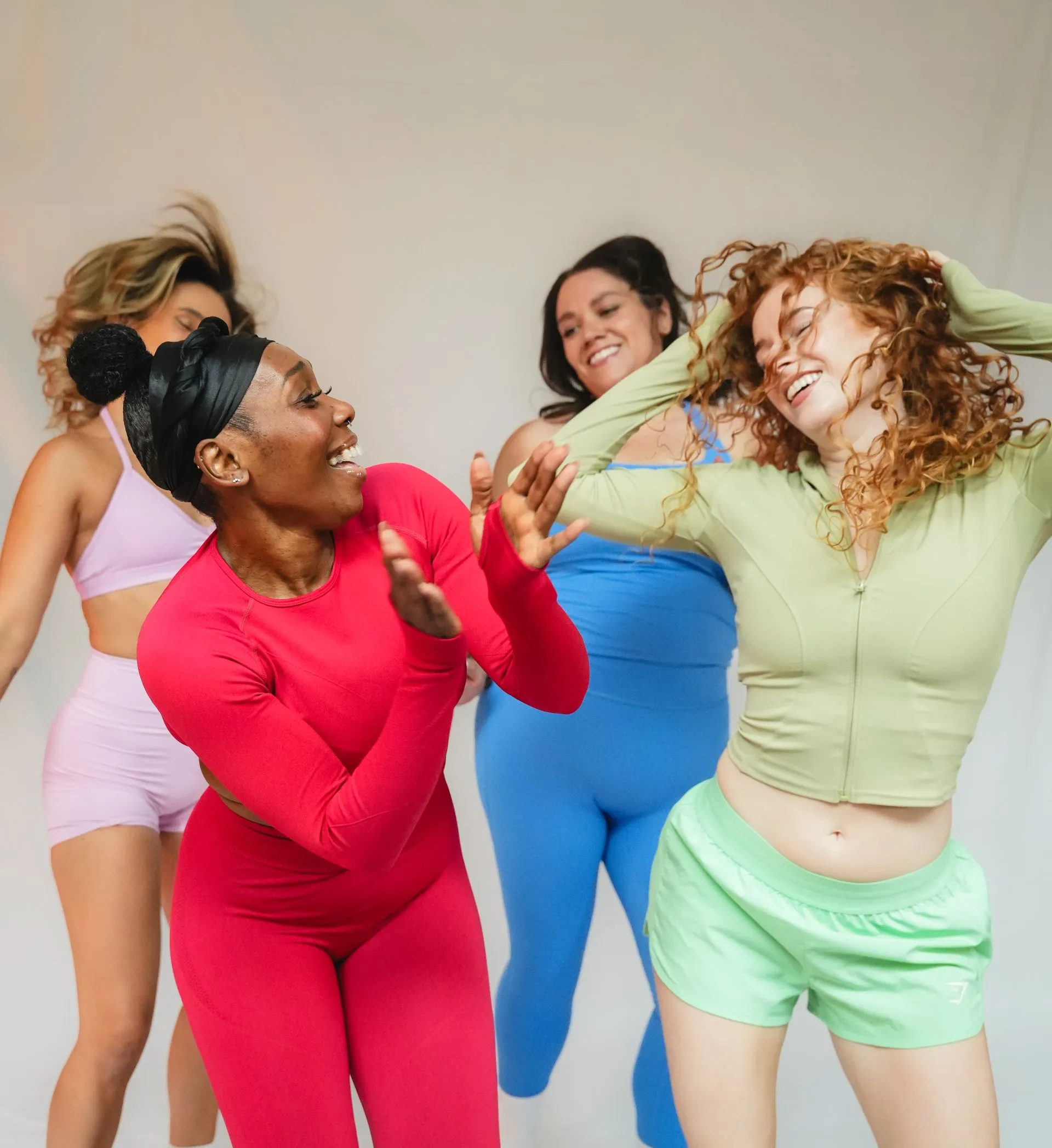 Four smiling women dancing and having fun in colourful activewear against a plain light background.
