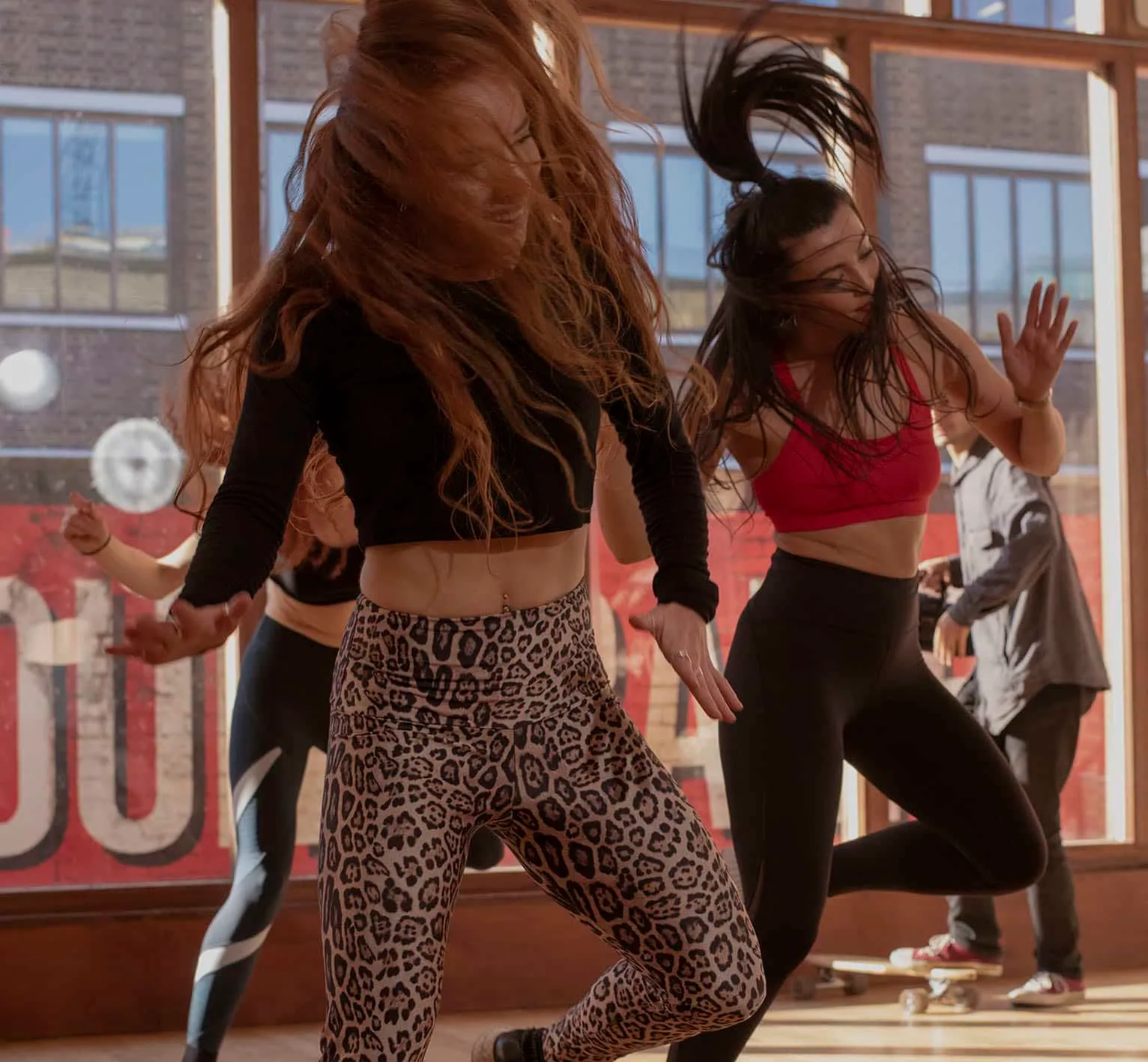 Two women dancing energetically in a sunlit studio with large windows