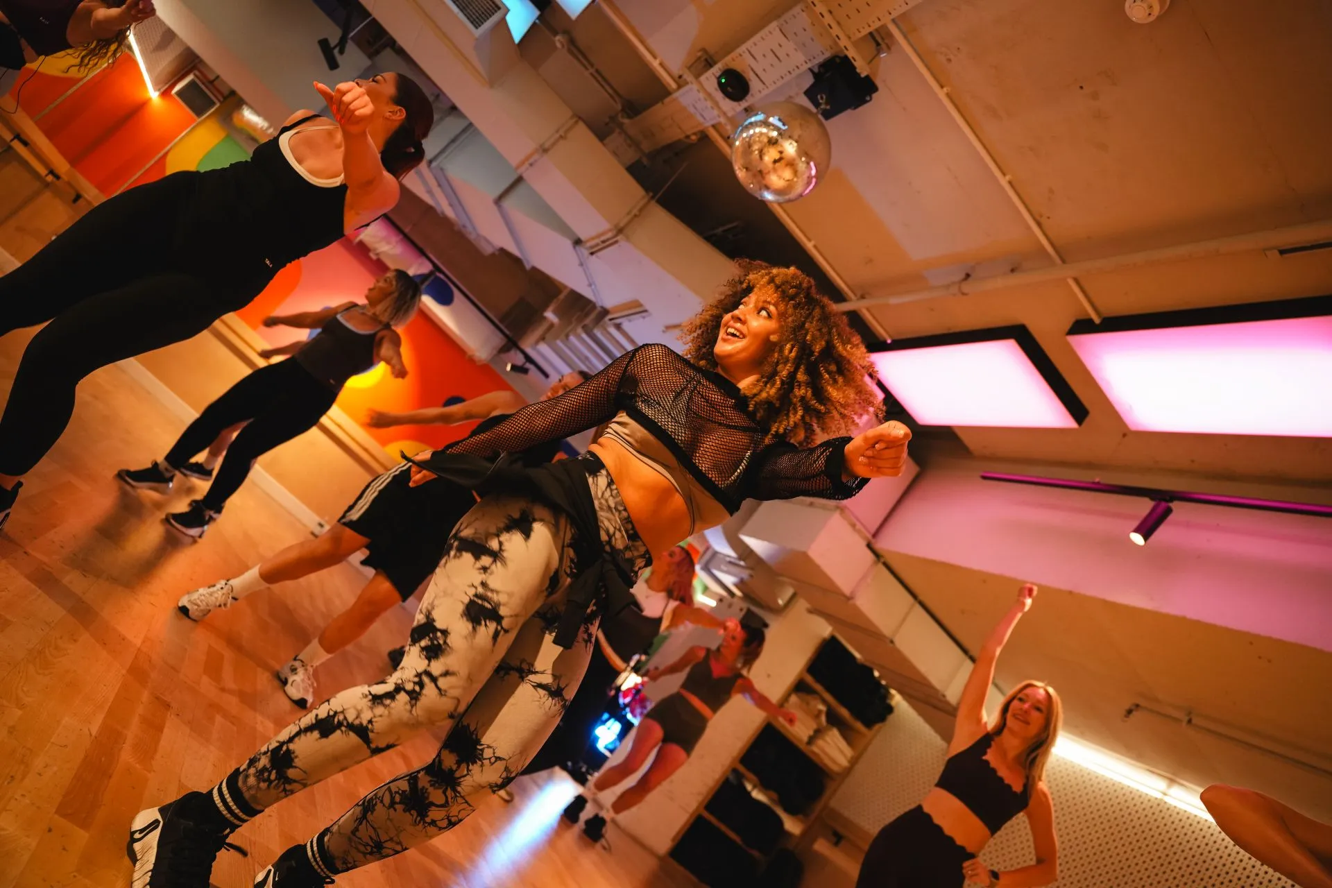 Group of women dancing energetically in a brightly lit dance studio with wooden floors, colourful walls, and ceiling lights, including a woman with curly hair wearing a black mesh top and patterned leggings in the foreground.