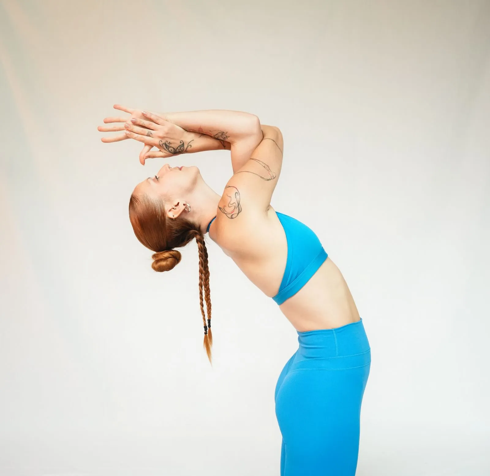 Woman with red braided hair and tattoos performing a standing yoga backbend pose, wearing a blue sports bra and leggings against a plain light background.