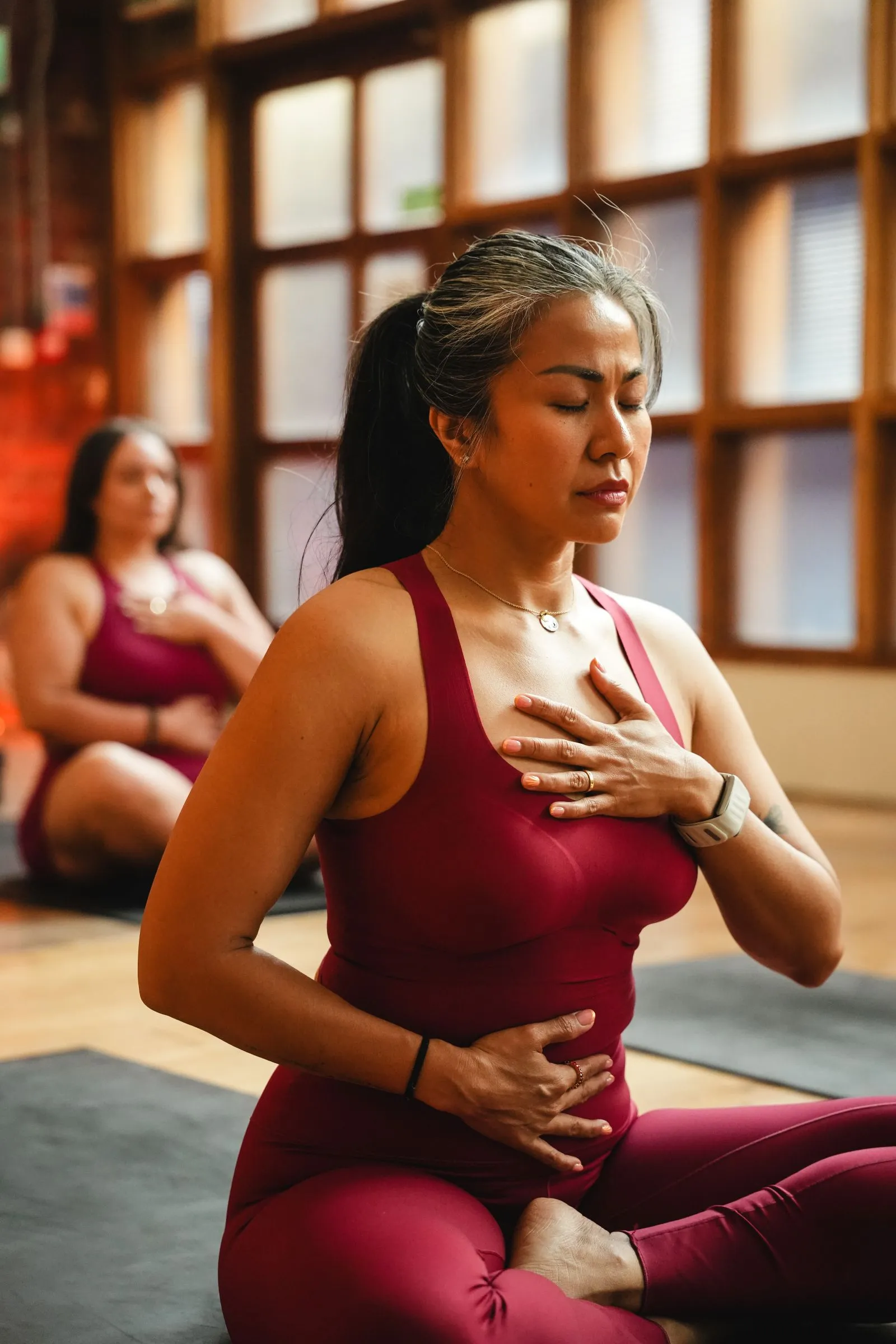 Two women in a yoga class sit cross-legged on mats, wearing matching maroon athletic outfits, with their eyes closed, meditating while placing one hand on their chest and the other on their abdomen in a tranquil room with large windows.