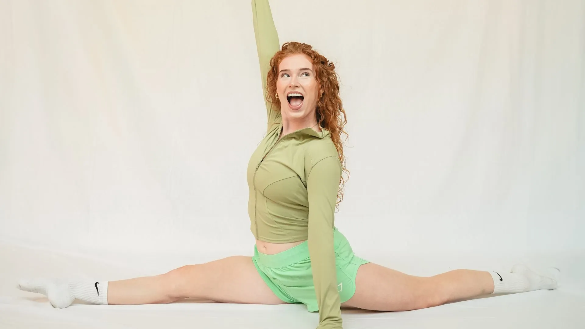 Smiling woman with red curly hair doing the splits with one arm raised