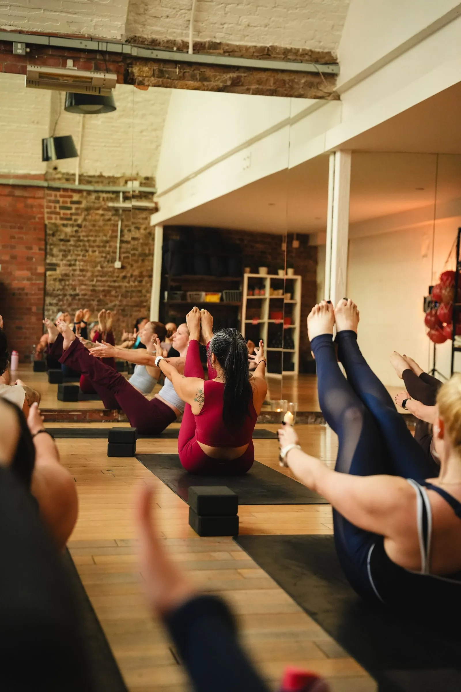 Class balancing in a seated V-sit on mats in a brick-walled studio with a mirrored wall