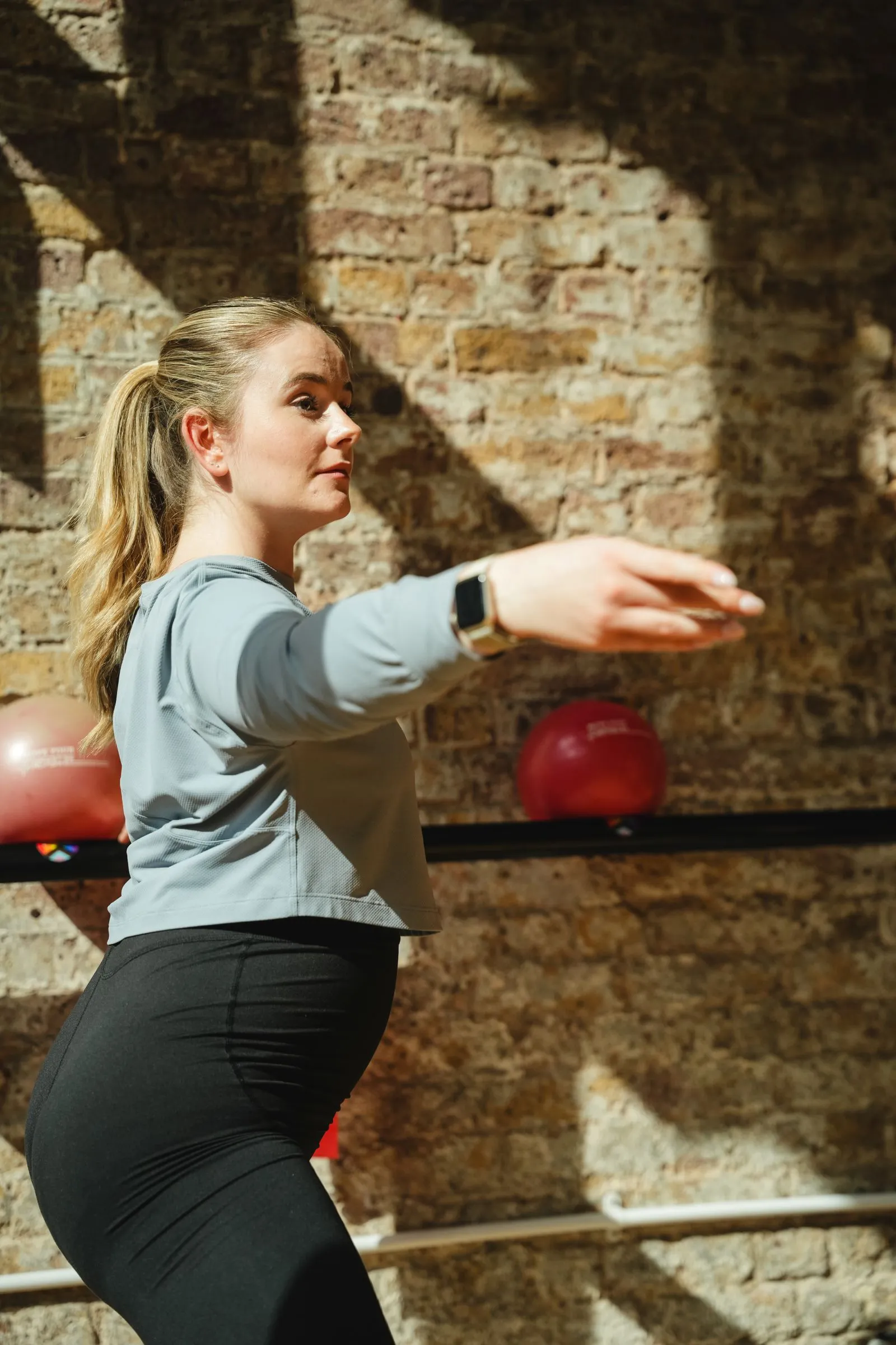 Pregnant woman with one arm extended at the barre in a sunlit brick-walled studio
