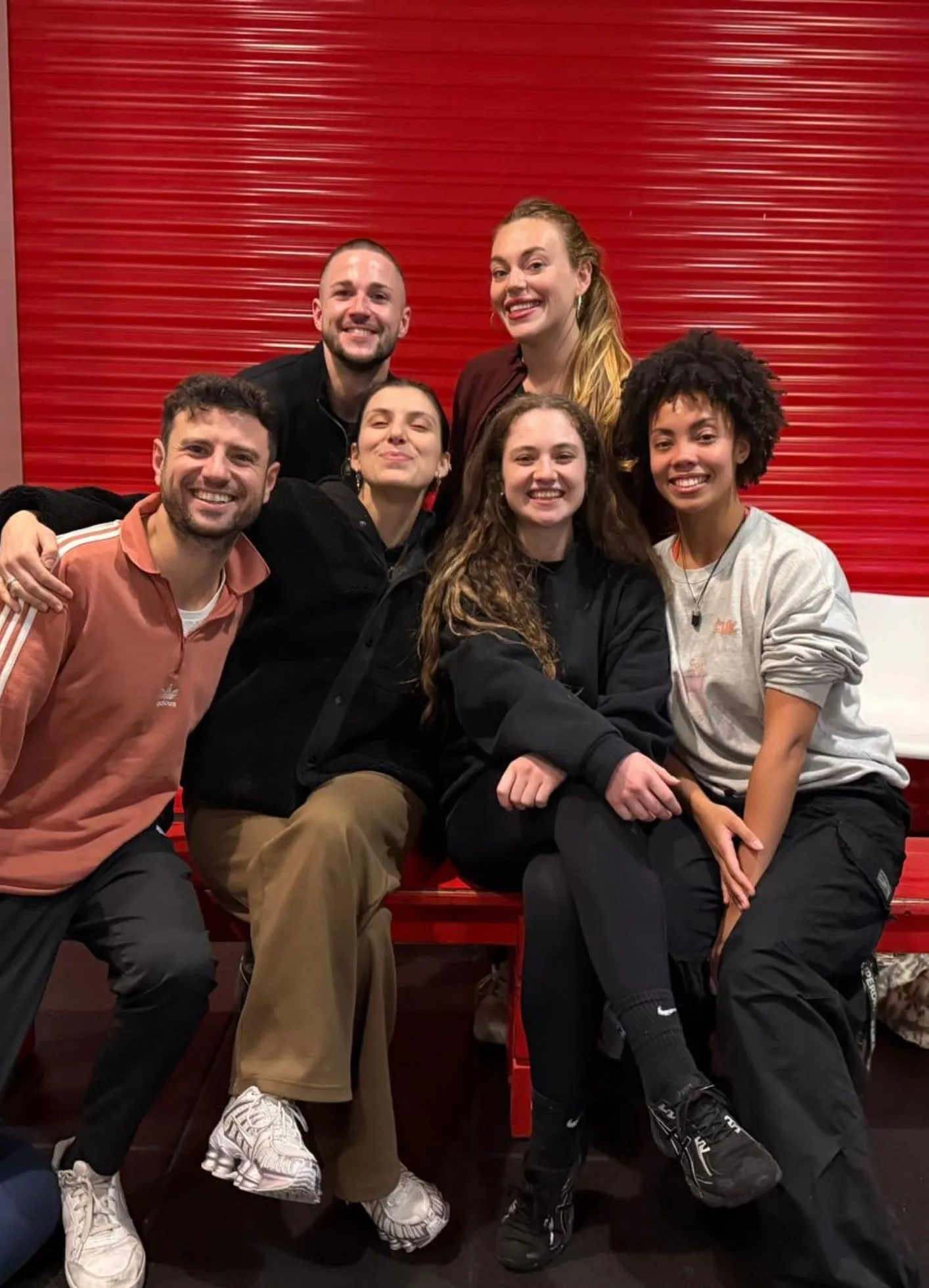 Group of seven smiling people sitting together in front of a red shutter