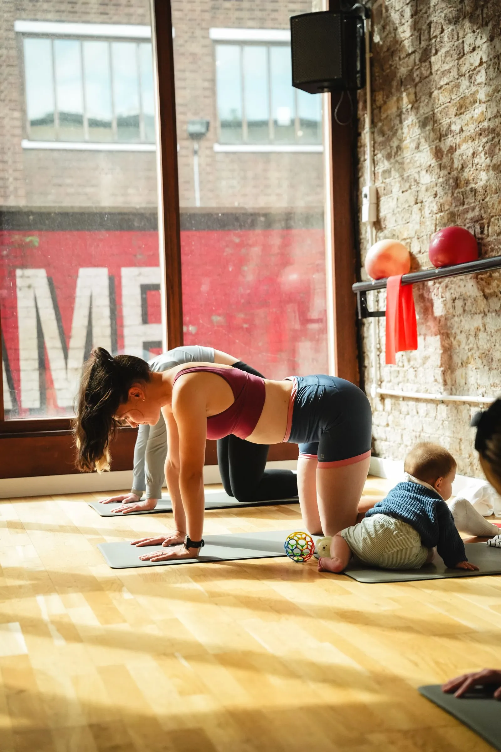Women doing a tabletop exercise on mats beside a baby in a sunlit studio