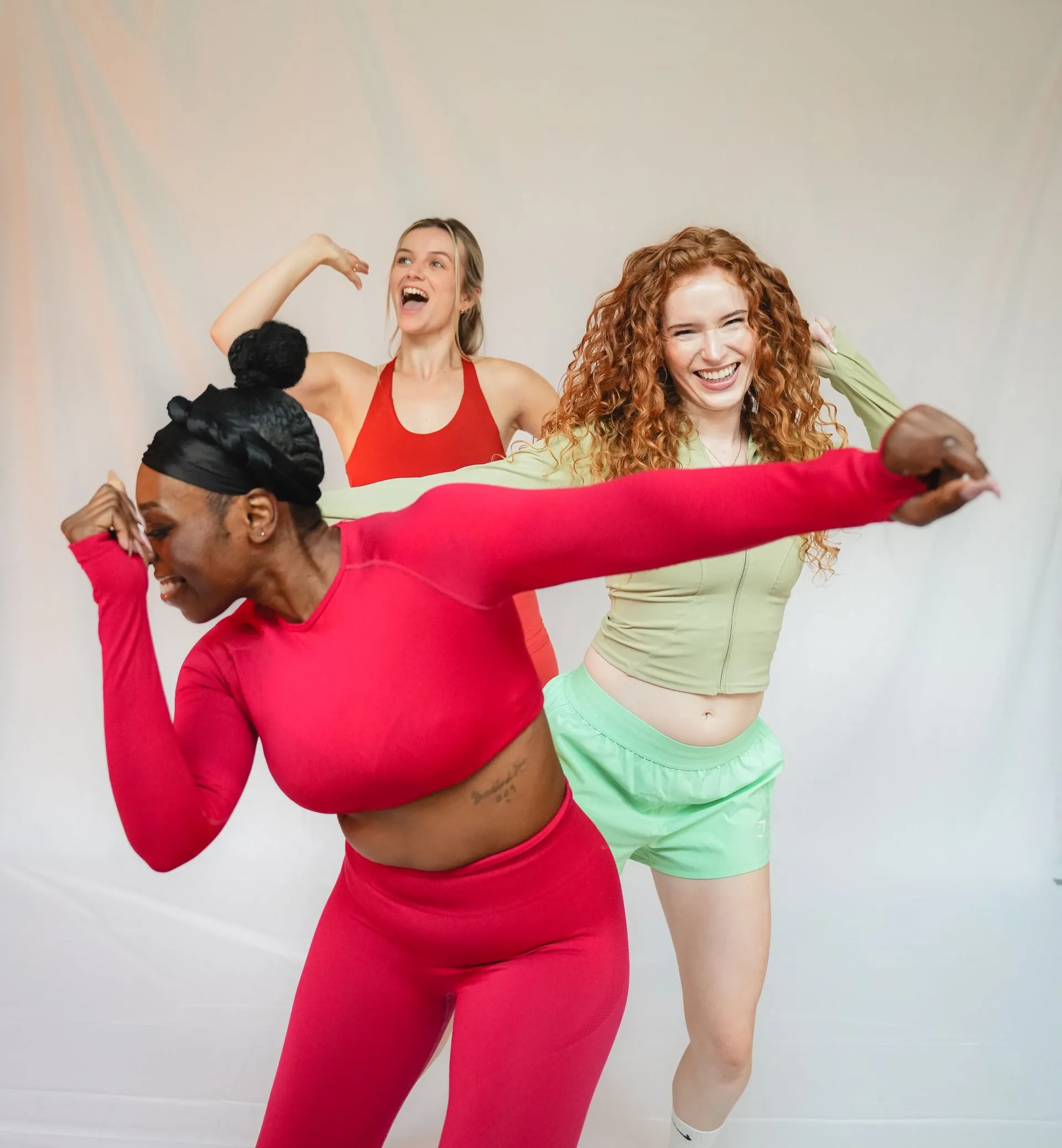 Three women laughing and dancing together in bright pink and green activewear