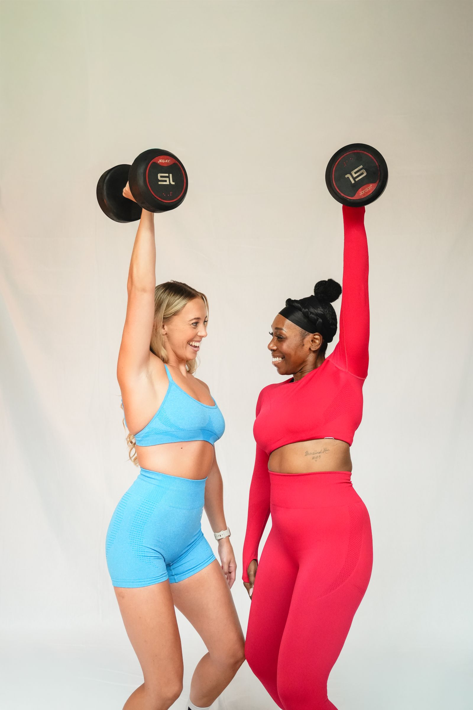 Two smiling women lifting dumbbells overhead in blue and red activewear