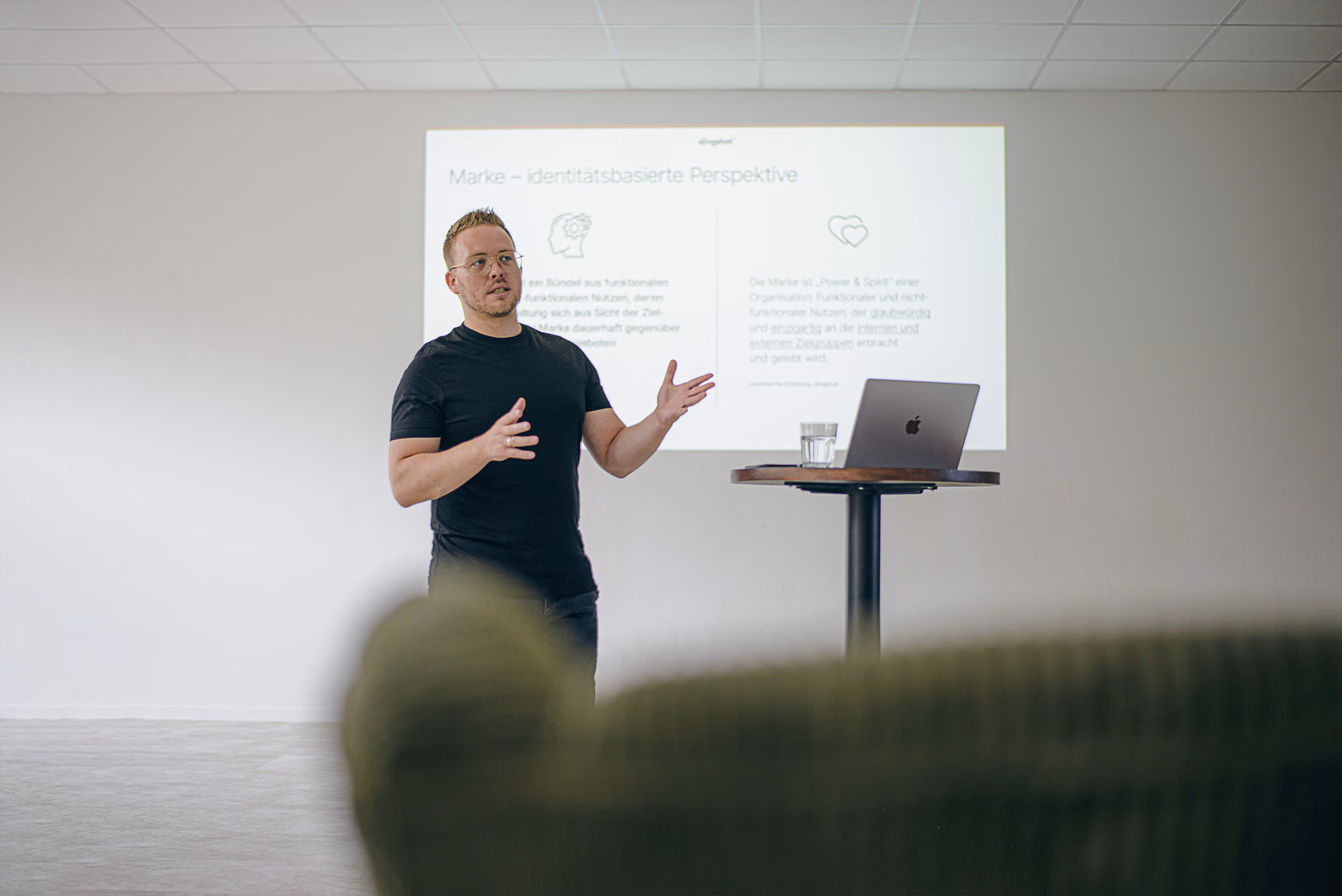 Stefan Klein, Inhaber von slingshot®, spricht gestikulierend neben einem Stehtisch mit Laptop vor einer Präsentation mit dem Titel ‚Marke – identitätsbasierte Perspektive‘.