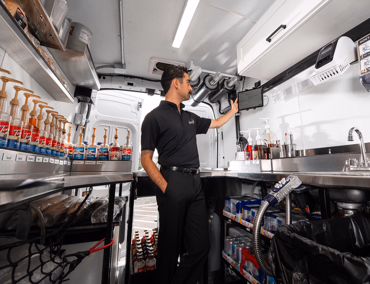 Man in black uniform stands inside a CoffeeNow van, touching a screen monitor mounted on the wall, surrounded by syrup bottles, drink supplies, and a sink.