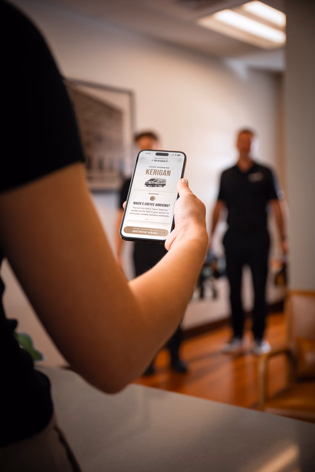 Person holding a smartphone displaying a food delivery app with a coffee order, two blurred figures standing in the background inside a room.