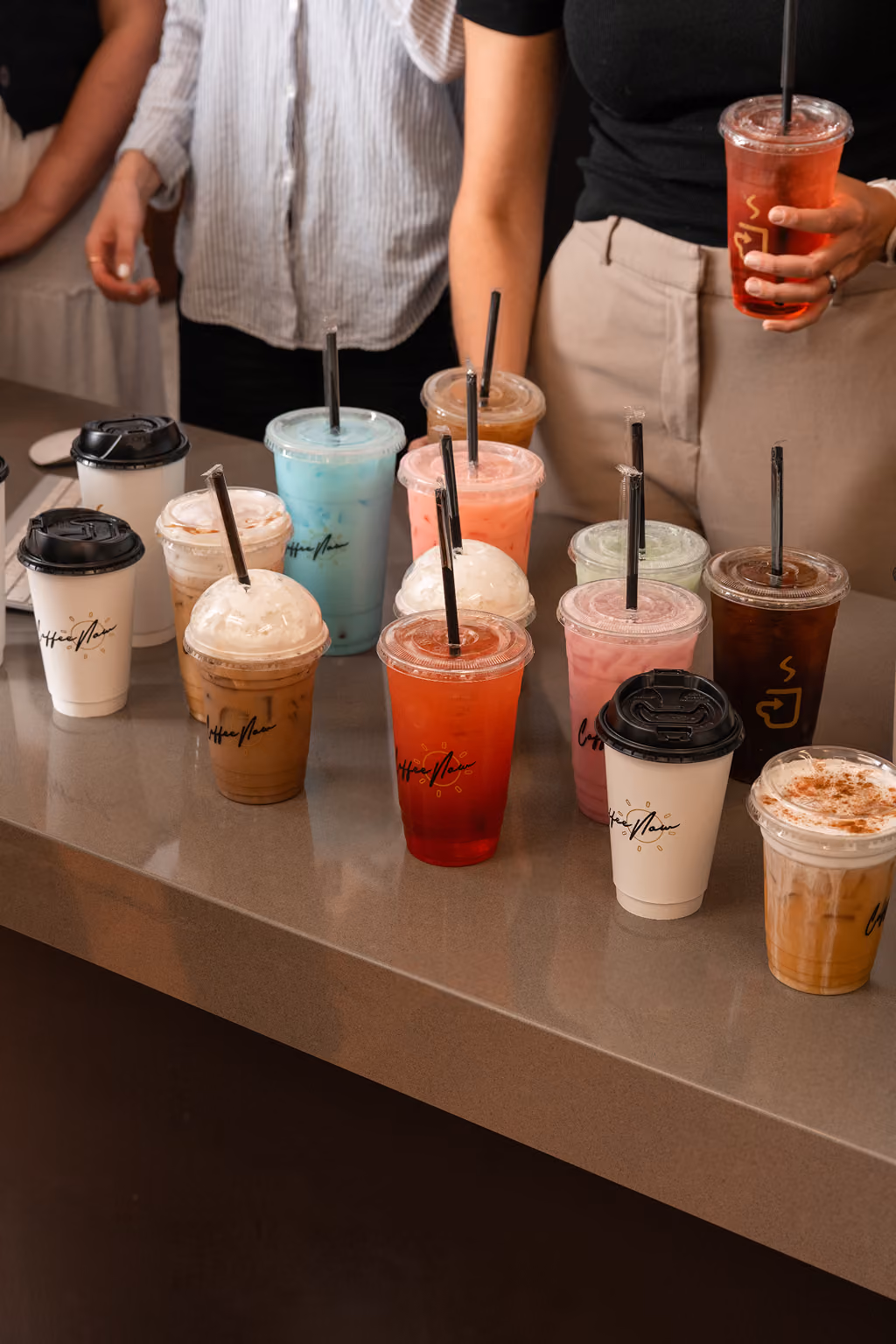 A variety of colorful iced and hot drinks in plastic and paper cups with straws arranged on a countertop, with two people standing behind them, one holding a red iced drink.