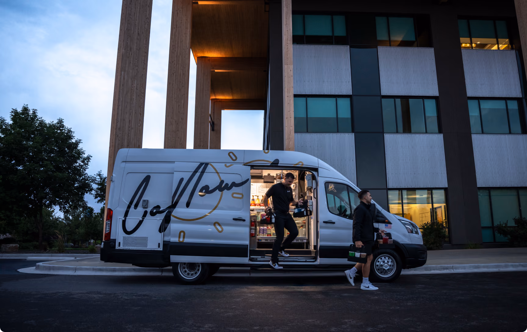CoffeeNow van parked outside an office building at dusk, with a barista handing off drinks