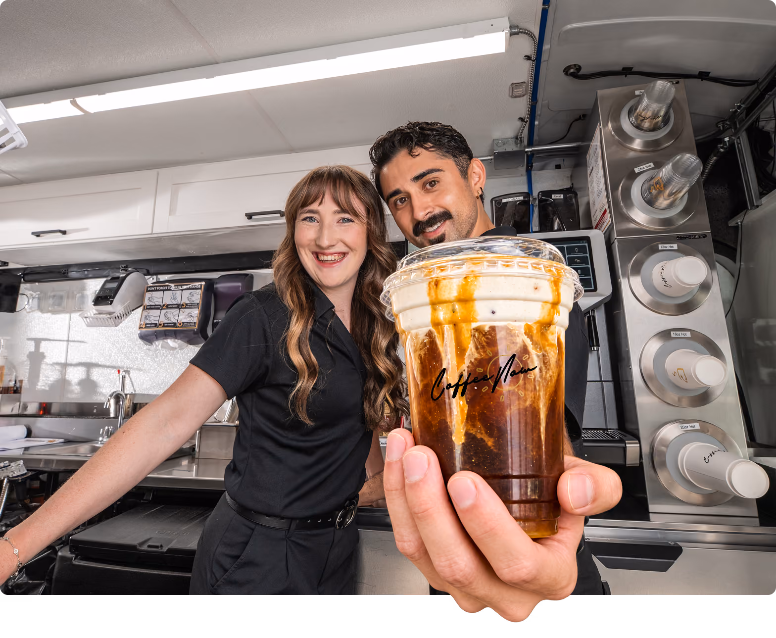 A smiling man and woman in black uniforms hold up a plastic cup of iced coffee topped with cream and caramel drizzle inside a coffee shop or food truck.