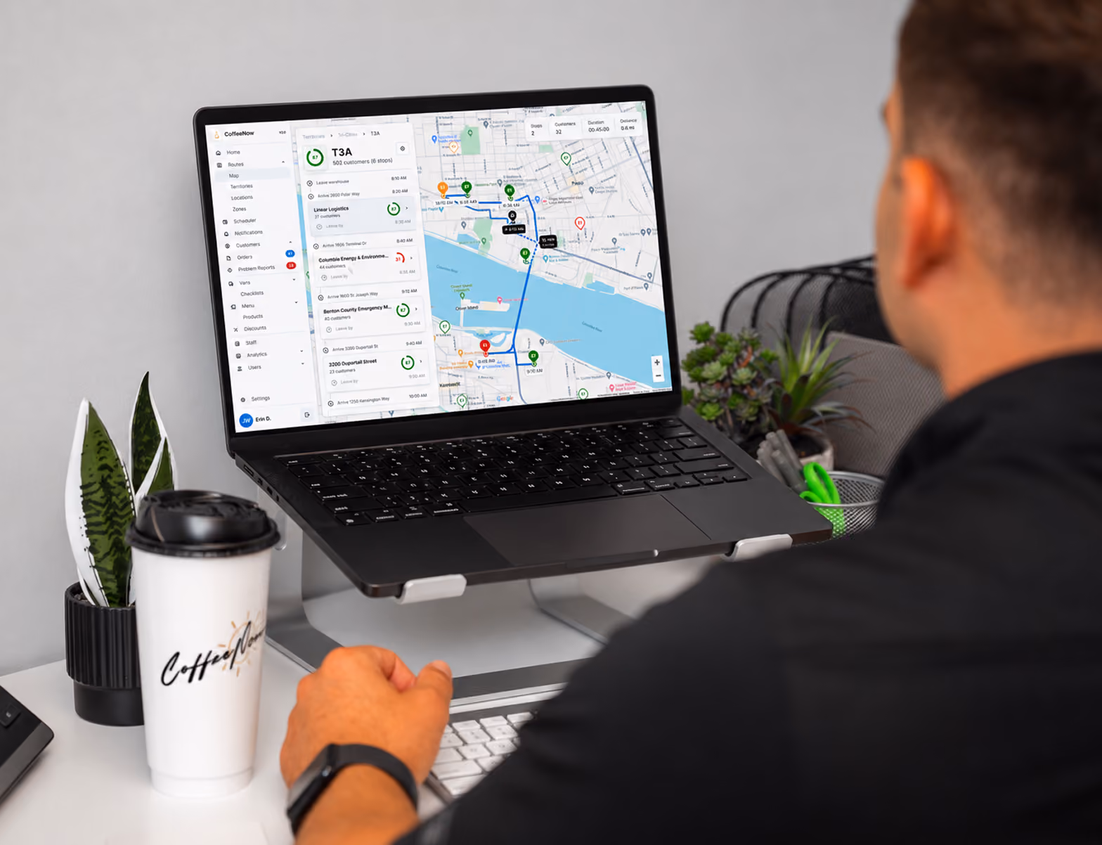 Person sitting at a desk using a laptop displaying a map with route navigation and delivery details in a logistics dashboard interface; desk features plants and a white coffee cup labeled Coffee Co.