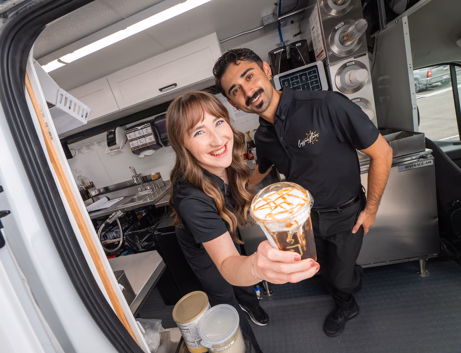 Two baristas inside a CoffeeNow van, smiling, with a woman handing a caramel-drizzled iced coffee towards the camera.