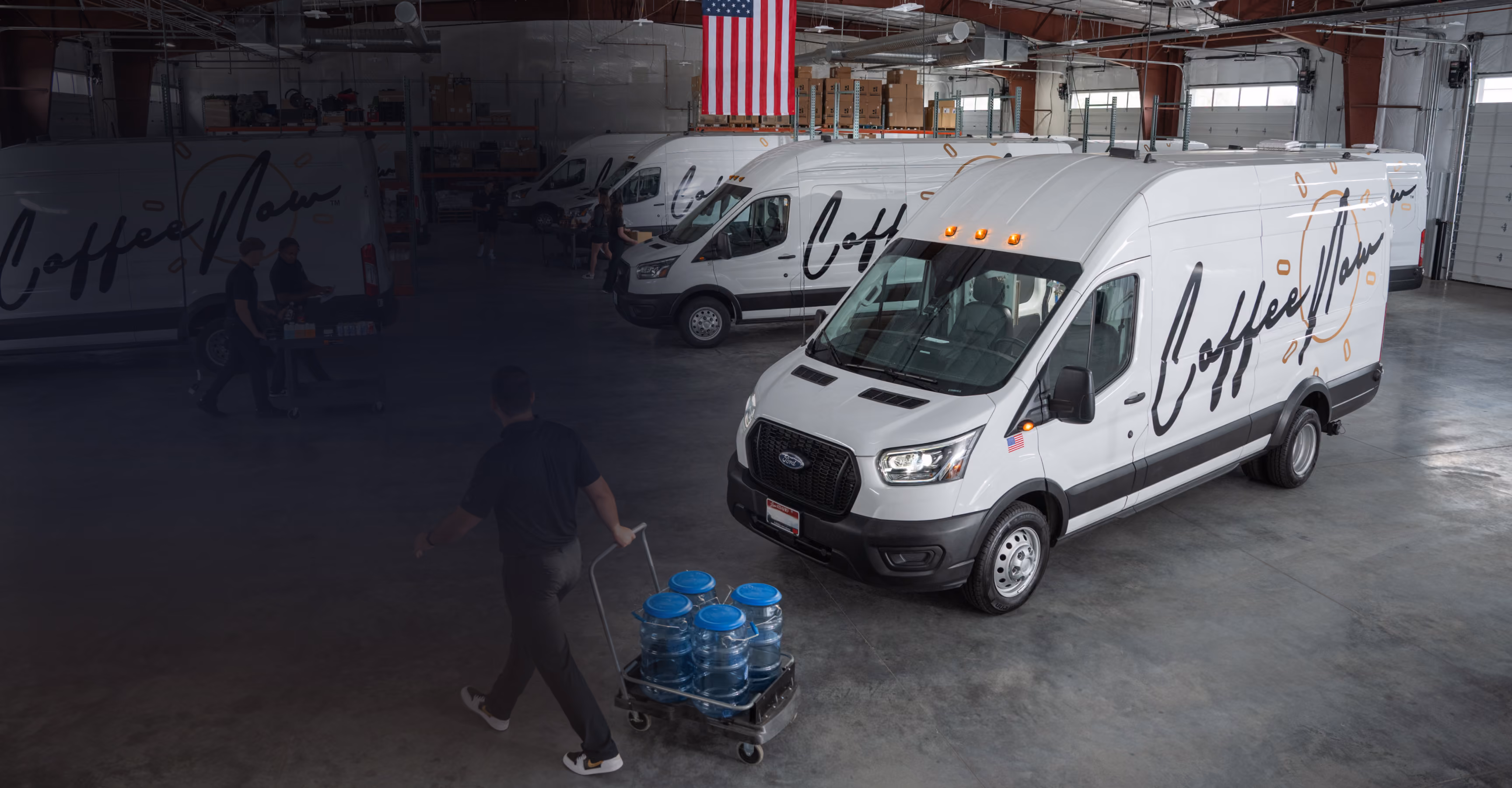 Warehouse interior with several white delivery vans lined up, each branded with the word 'Coffee Man' and images of coffee beans; a person is pushing a cart loaded with blue water jugs across the concrete floor.