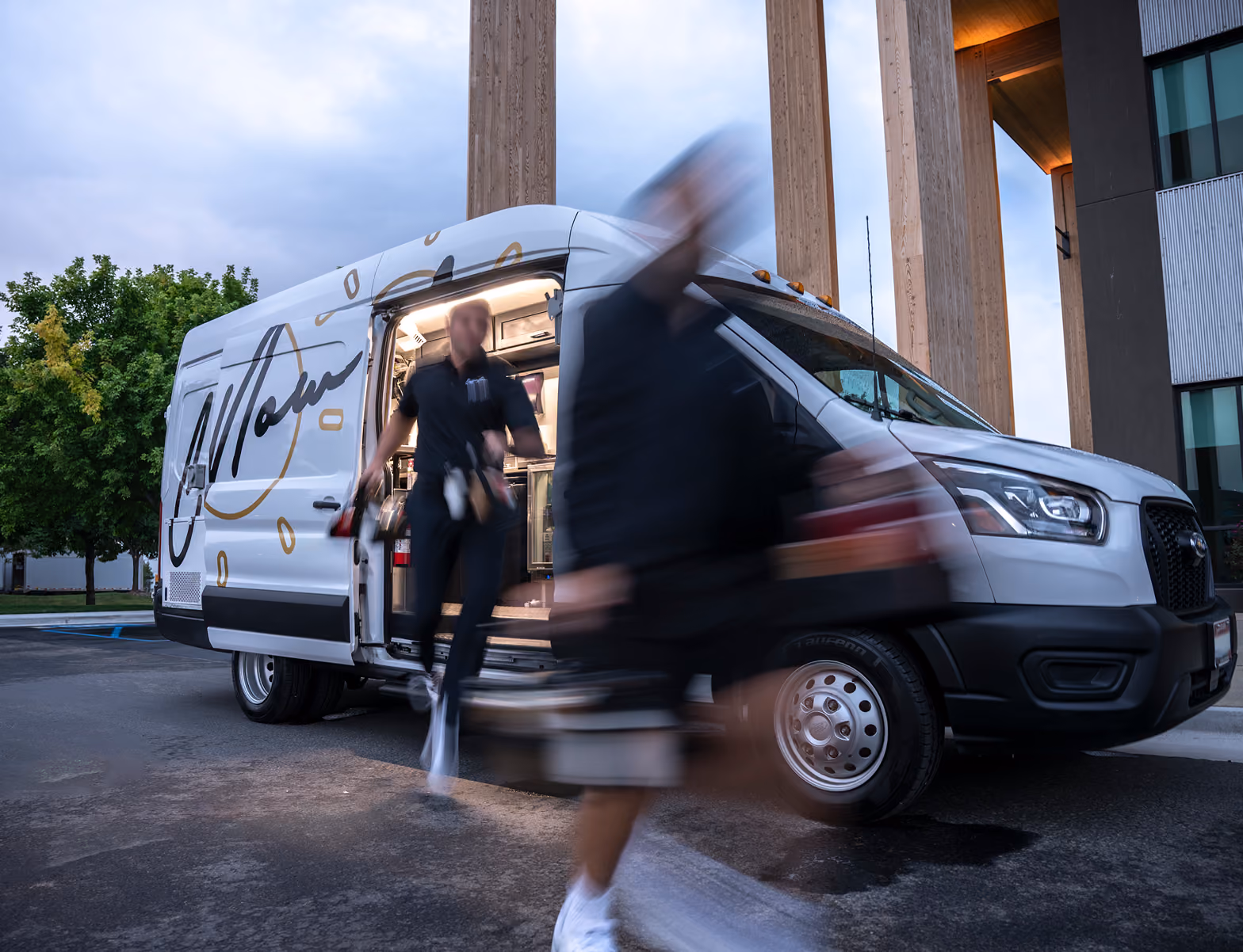 White CoffeeNow delivery van with a stylized script logo parked outside a building with tall columns, a person exits the van and another person carrying a box is walking in front, both figures are blurred from motion.