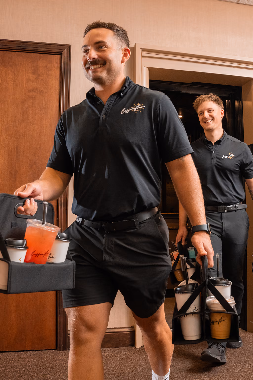 Two smiling men wearing black shirts and shorts carrying trays with multiple iced coffee drinks in a hallway with wooden and beige walls.
