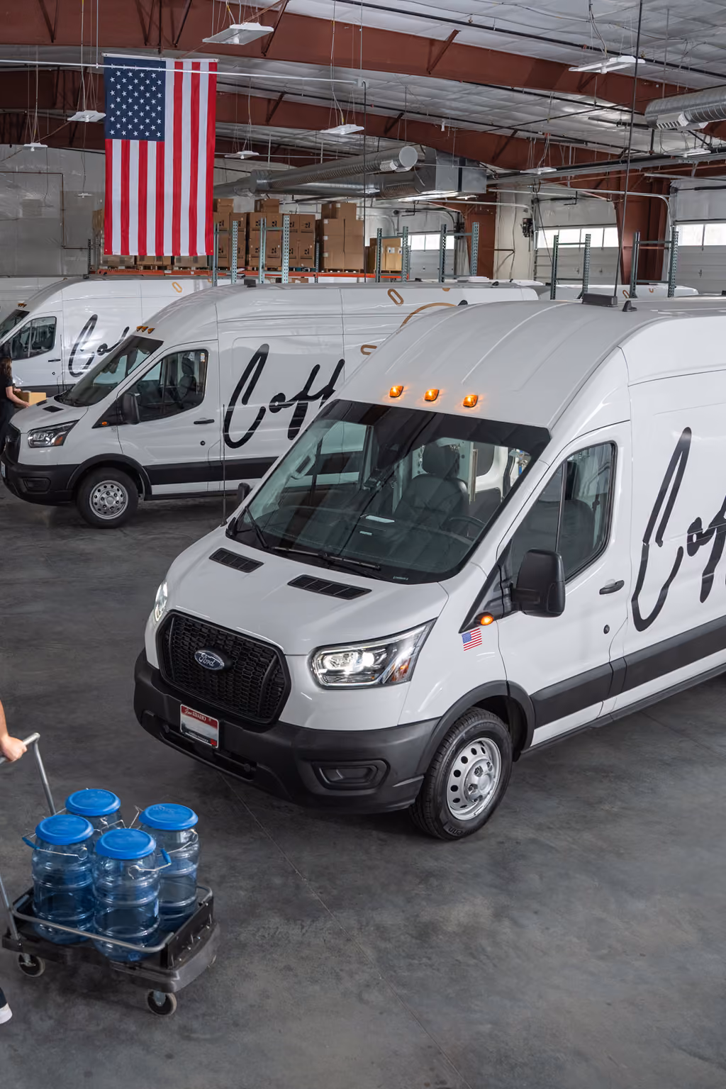 Three white CoffeeNow vans with black script logos parked inside a warehouse with an American flag hanging from the ceiling, and a person pushing a cart loaded with large water bottles on the left.
