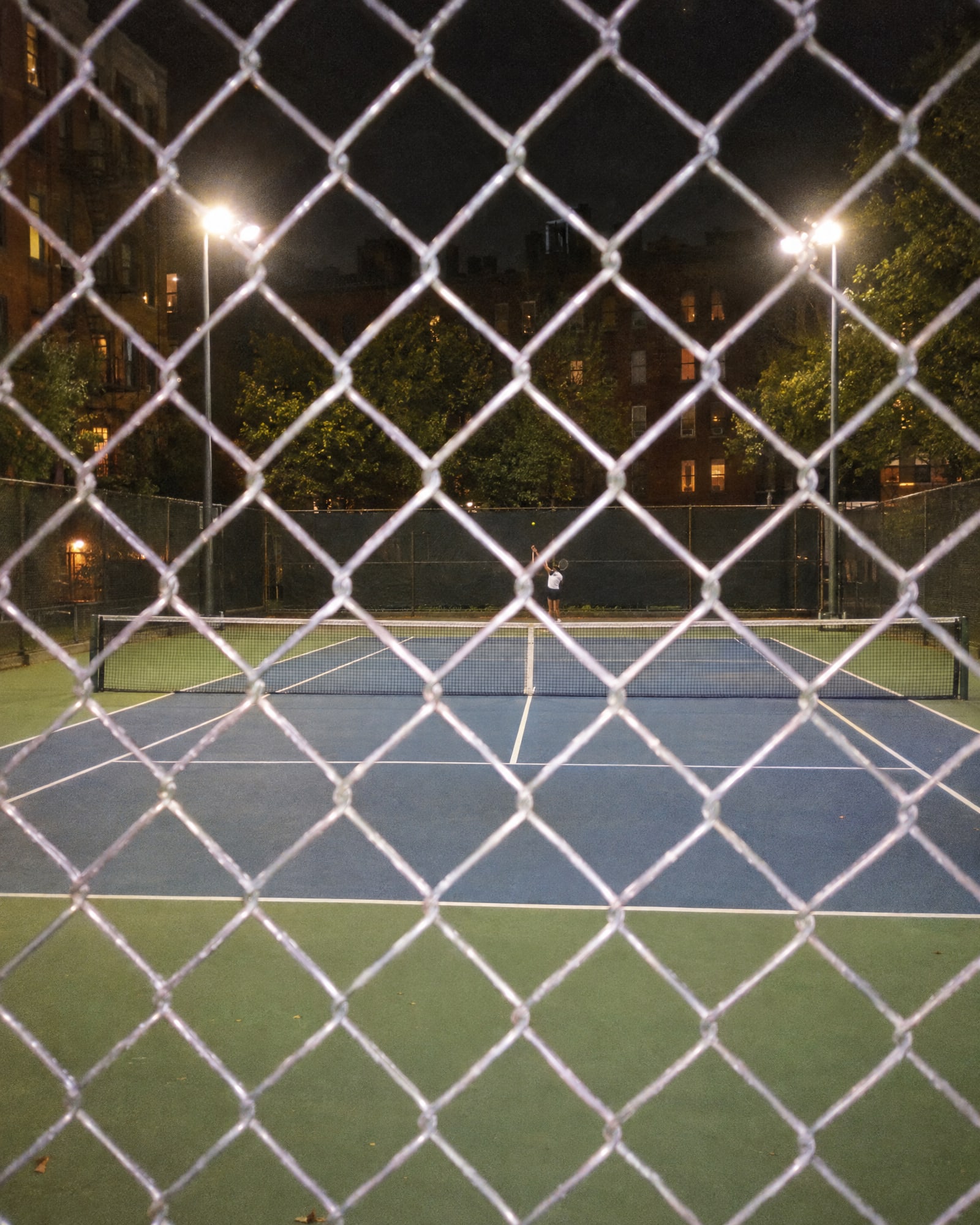 A lit tennis court at night through a chain-link fence, one player at the far baseline.