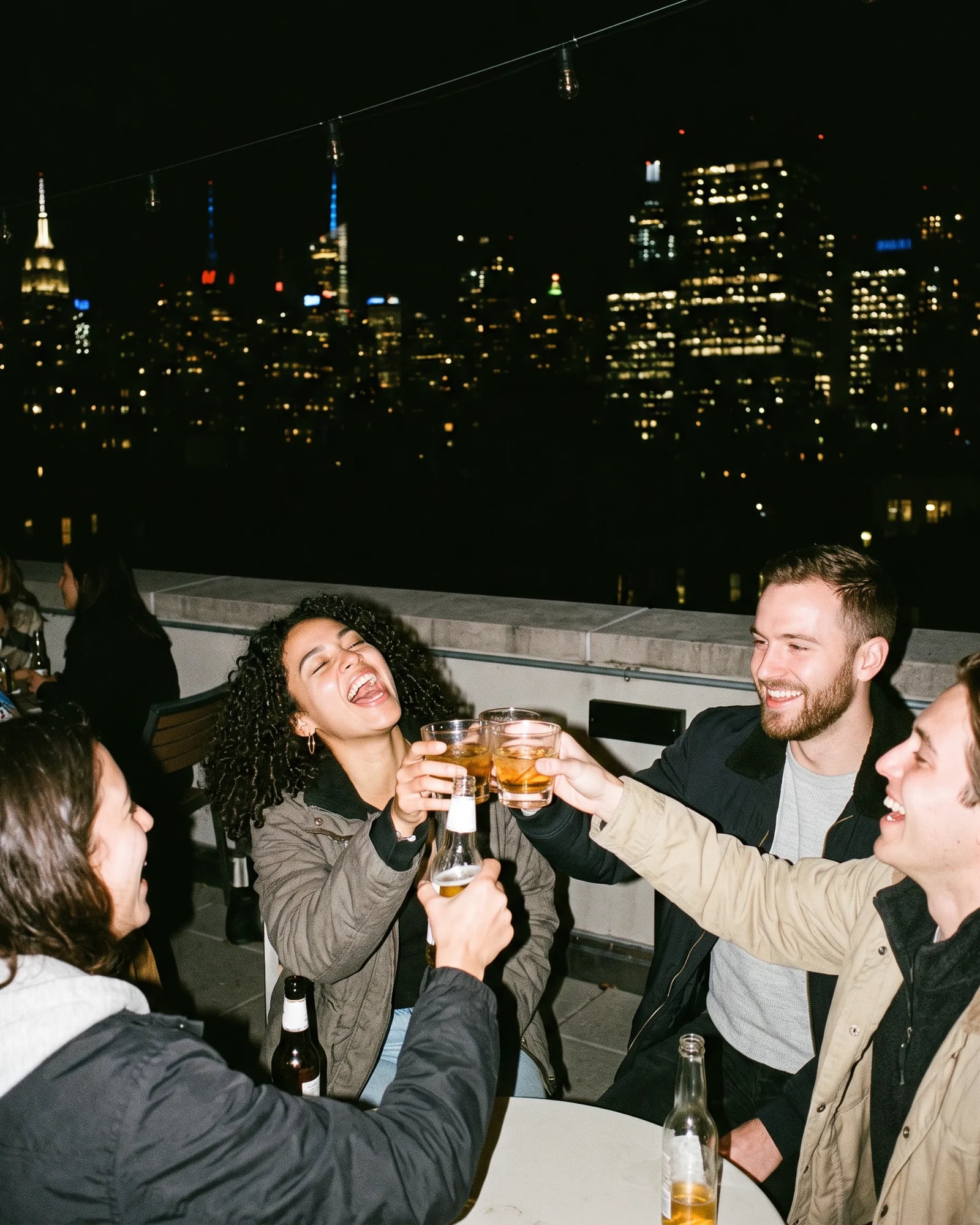 Friends toasting with drinks on a rooftop, the skyline lit behind them.
