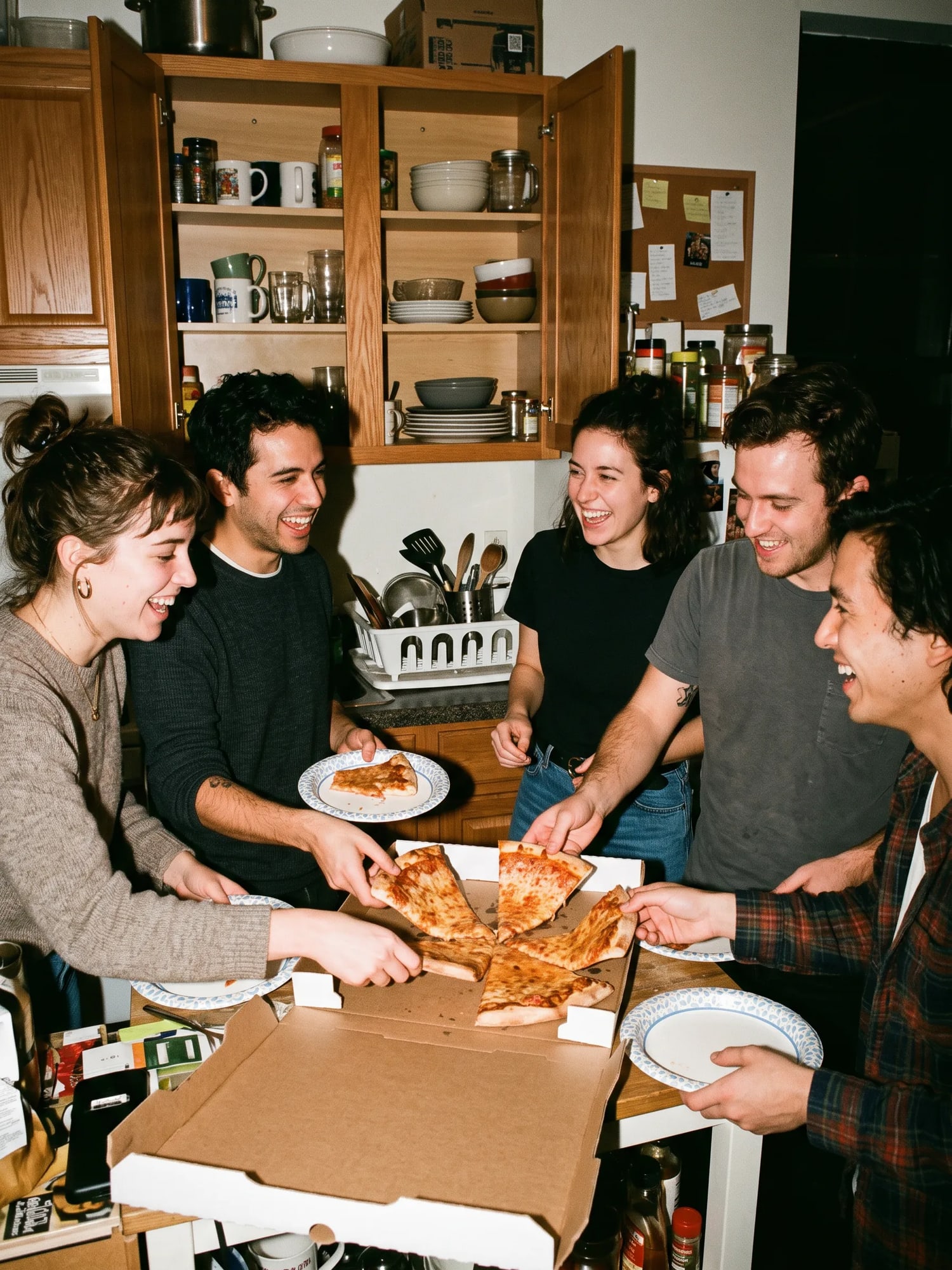 Friends reaching for slices of pizza in a small apartment kitchen.