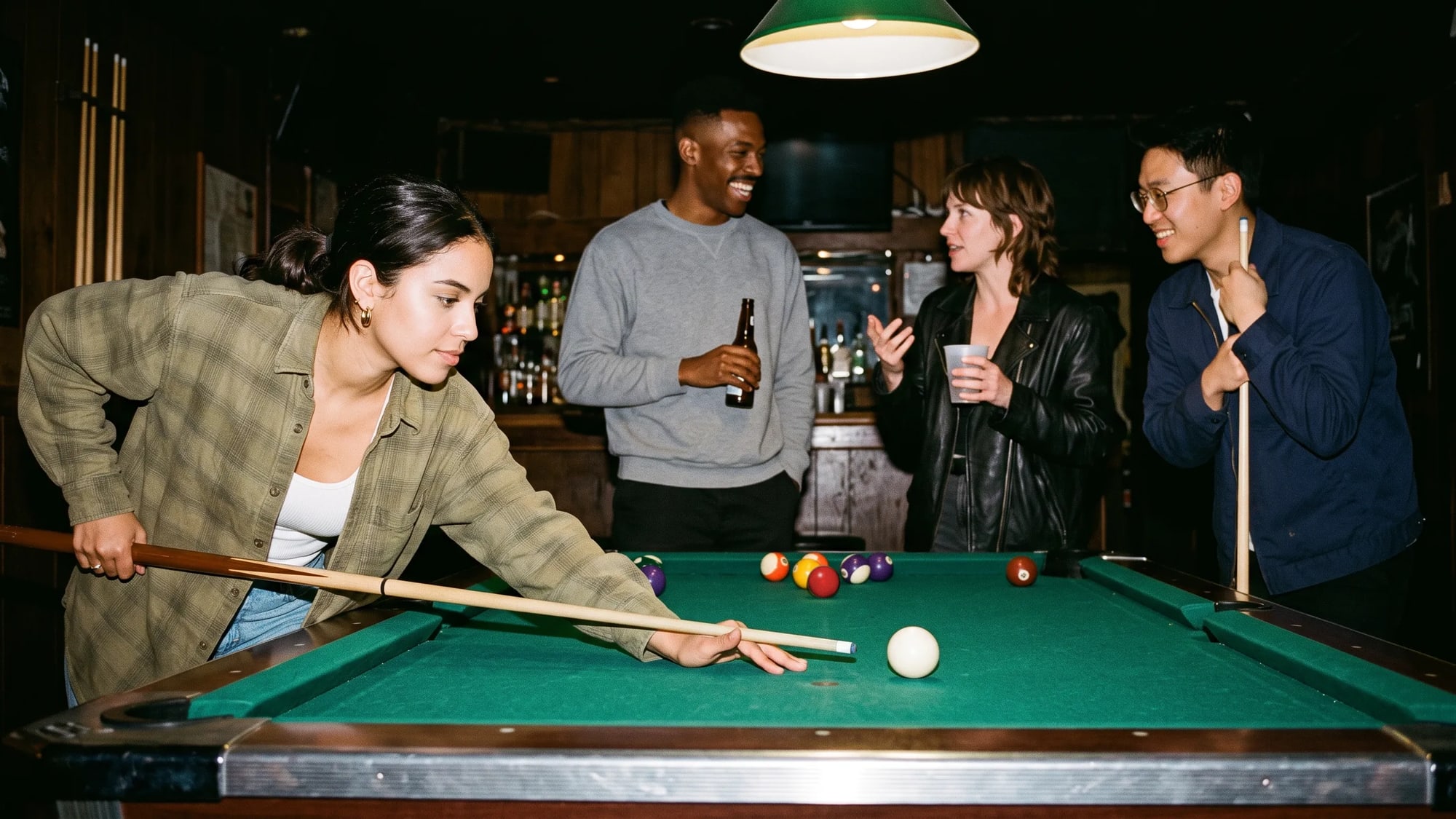 A young woman leans over a pool table to line up a shot on the cue ball while three friends talk and laugh behind the table in a dim bar.