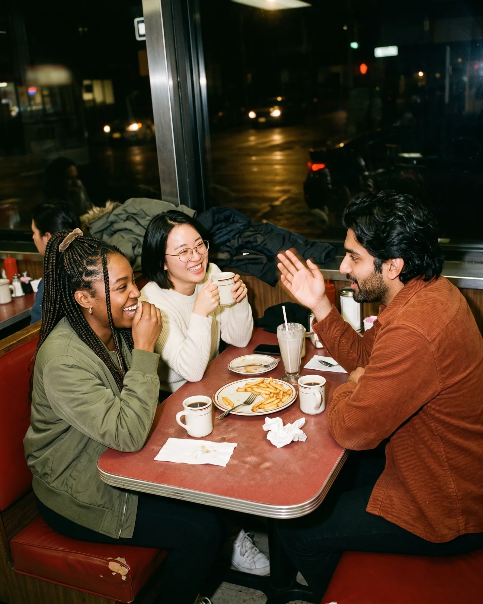 Three friends laugh together in a red diner booth at night while one of them tells a story over coffee and fries.
