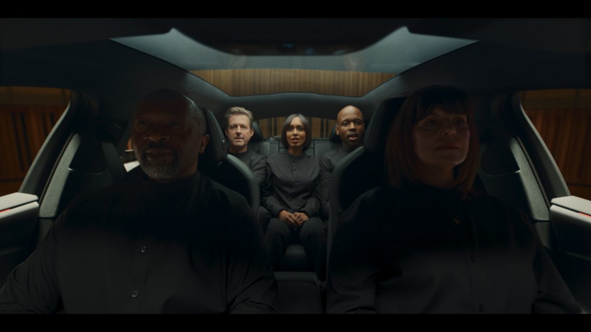 BBH ad still: Five choir members dressed in black sit inside an Audi A6 Avant e-tron, illuminated by soft natural light through the panoramic sunroof, with wood-panelled concert hall walls faintly visible outside the windows.